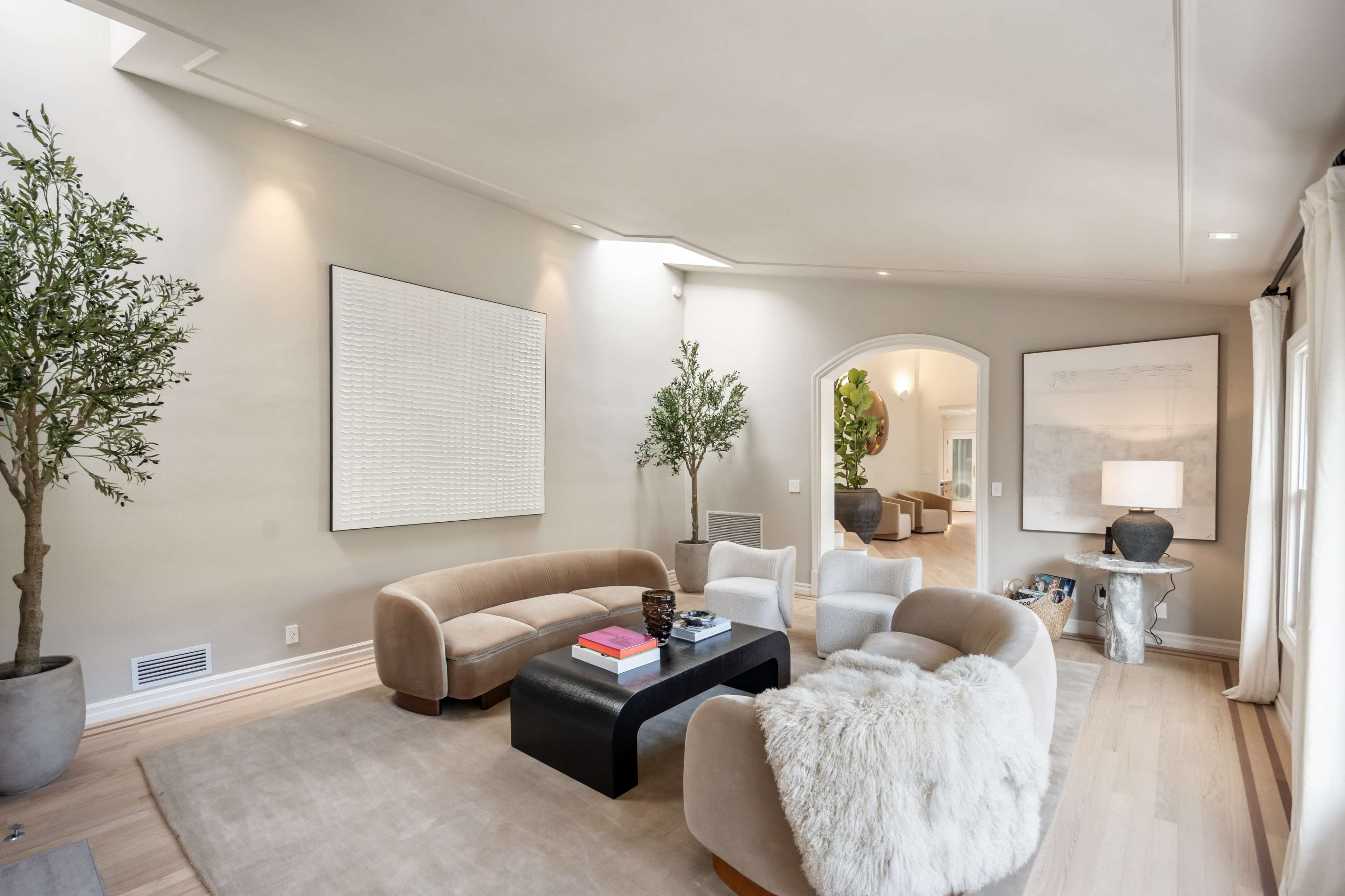 The image shows a modern living room with curved furniture, a textured wall art piece, and large windows letting in natural light.