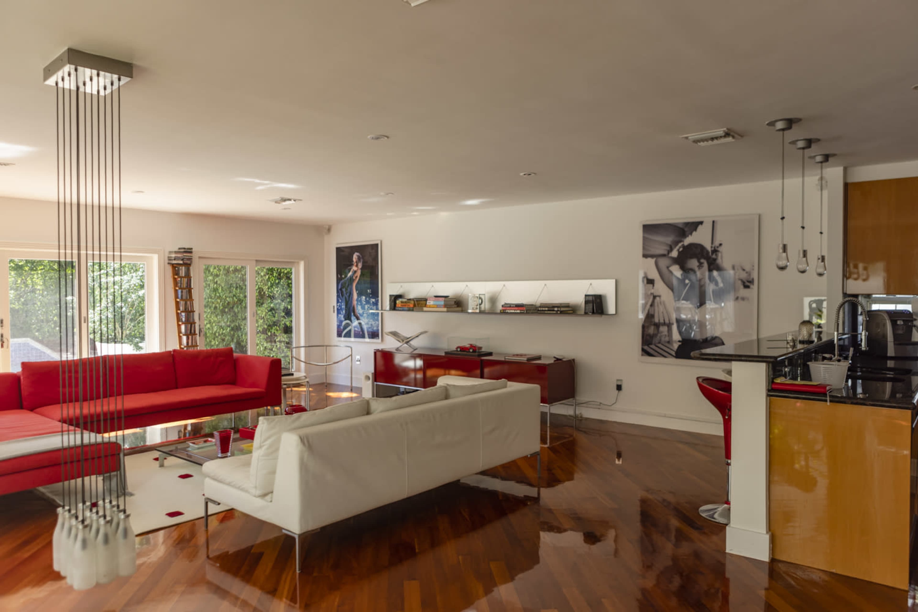 The image shows a modern living room with red and white furniture, hardwood floors, large windows, and a sleek kitchen area visible in the background.
