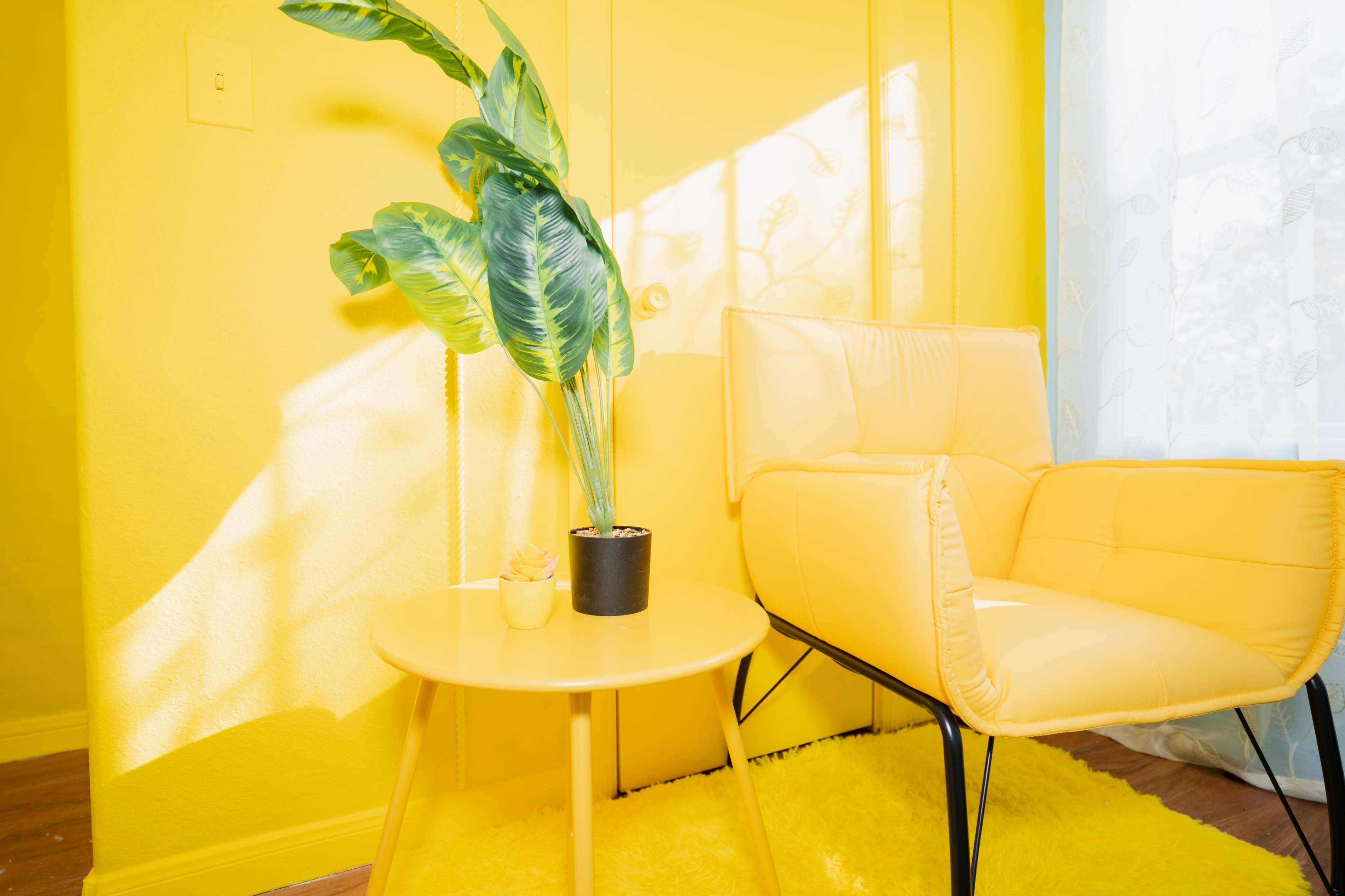 A bright yellow room features a yellow armchair next to a round table, with a potted plant on the table and sunlight casting shadows on the walls.