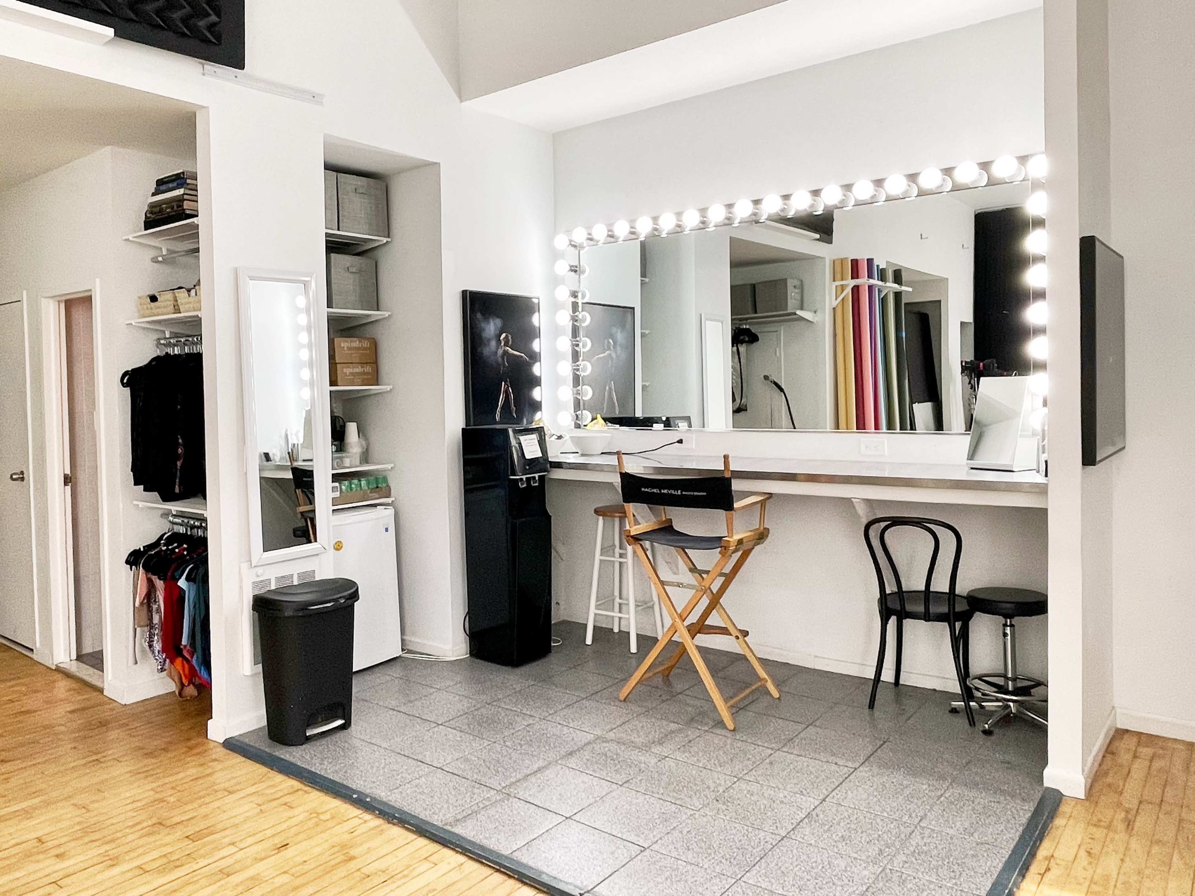 A well-lit dressing room with a large mirror surrounded by lights, a chair, and storage shelves.
