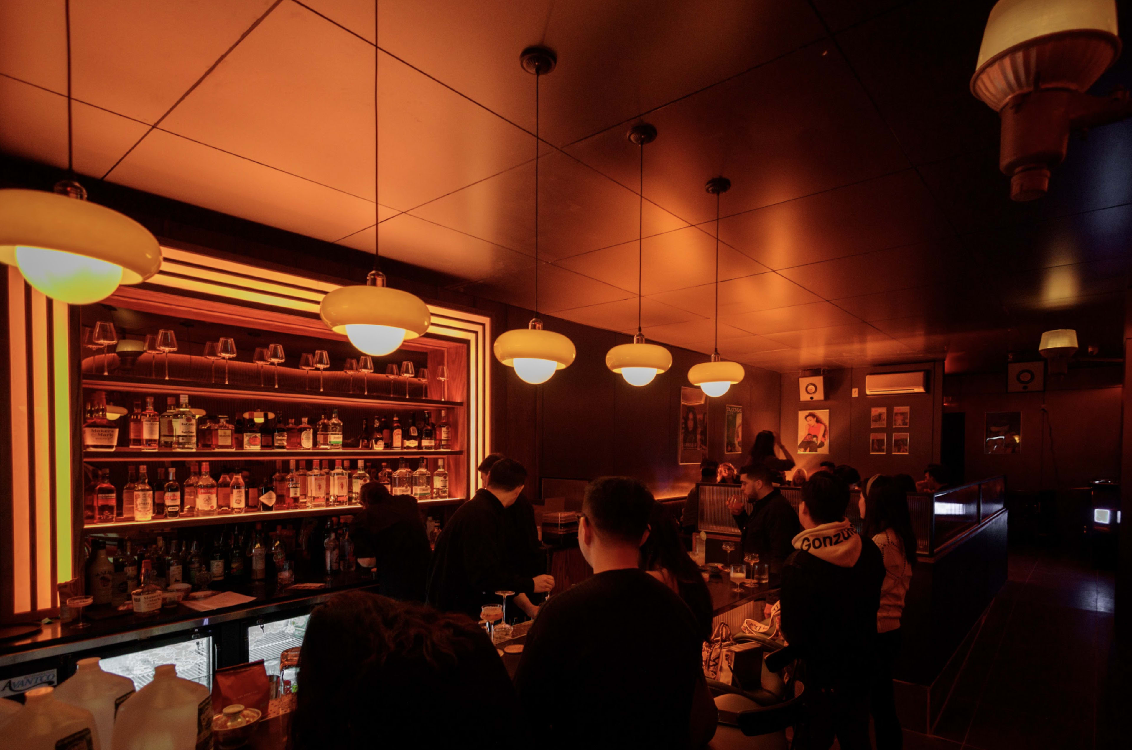 A bar scene features patrons interacting at a counter illuminated by warm orange neon lights and vintage-style pendant lamps, with shelves displaying various bottles in the background.