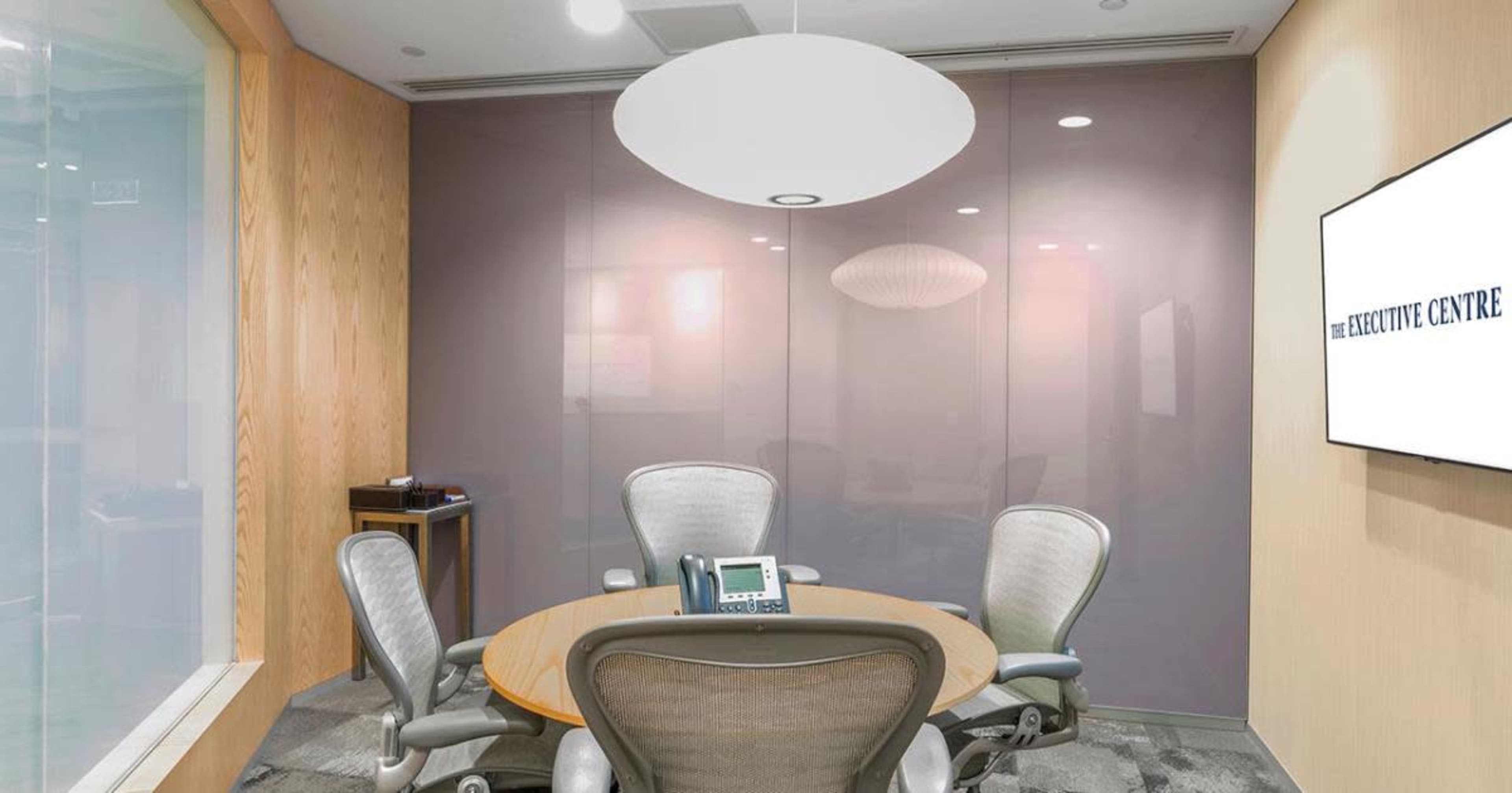 A modern conference room features a round wooden table and ergonomic chairs, with glass walls and a wall-mounted screen displaying "THE EXECUTIVE CENTRE."