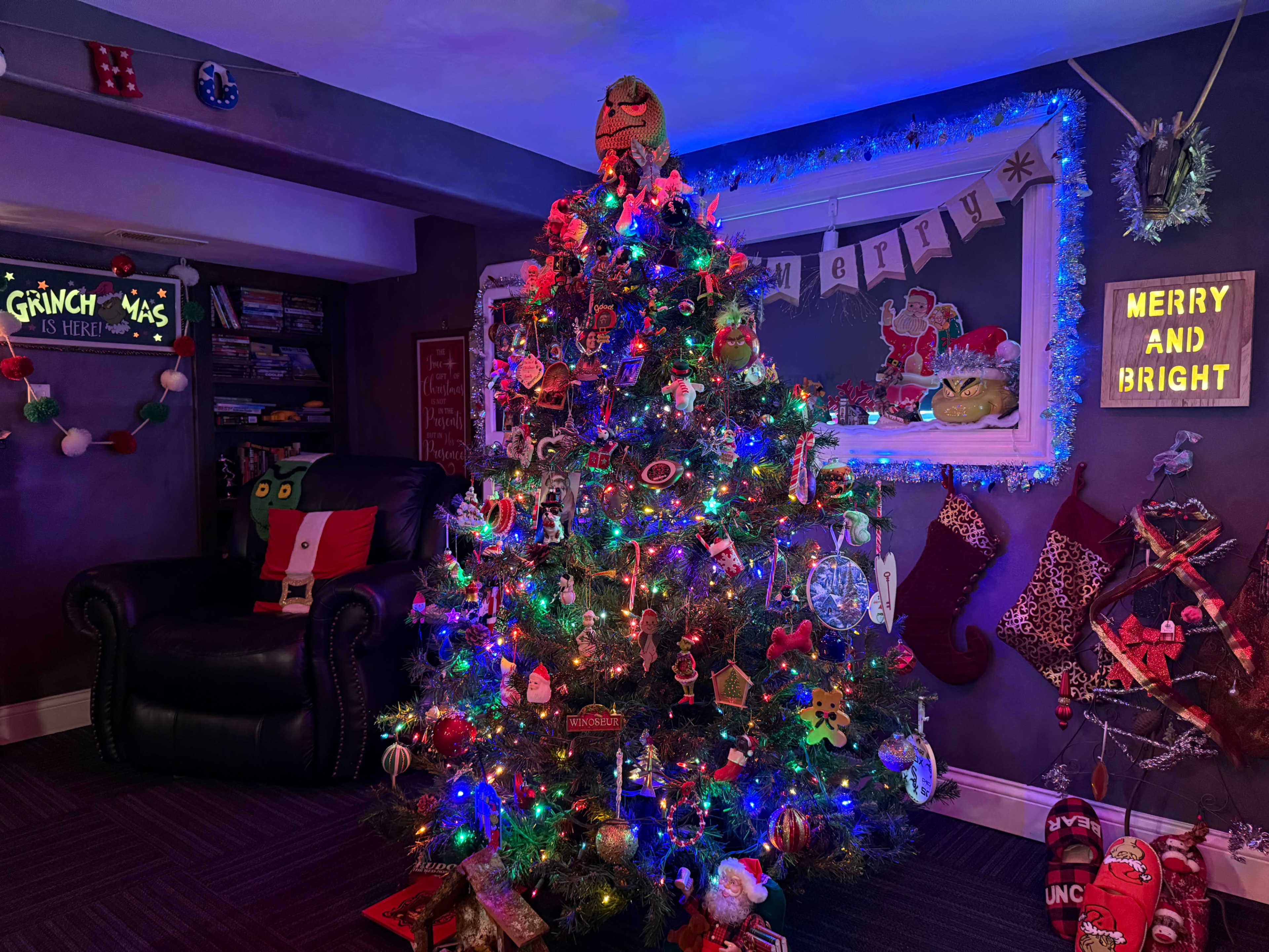 A brightly decorated Christmas tree adorned with colorful lights and ornaments stands in a cozy room featuring holiday-themed decorations and a leather chair.