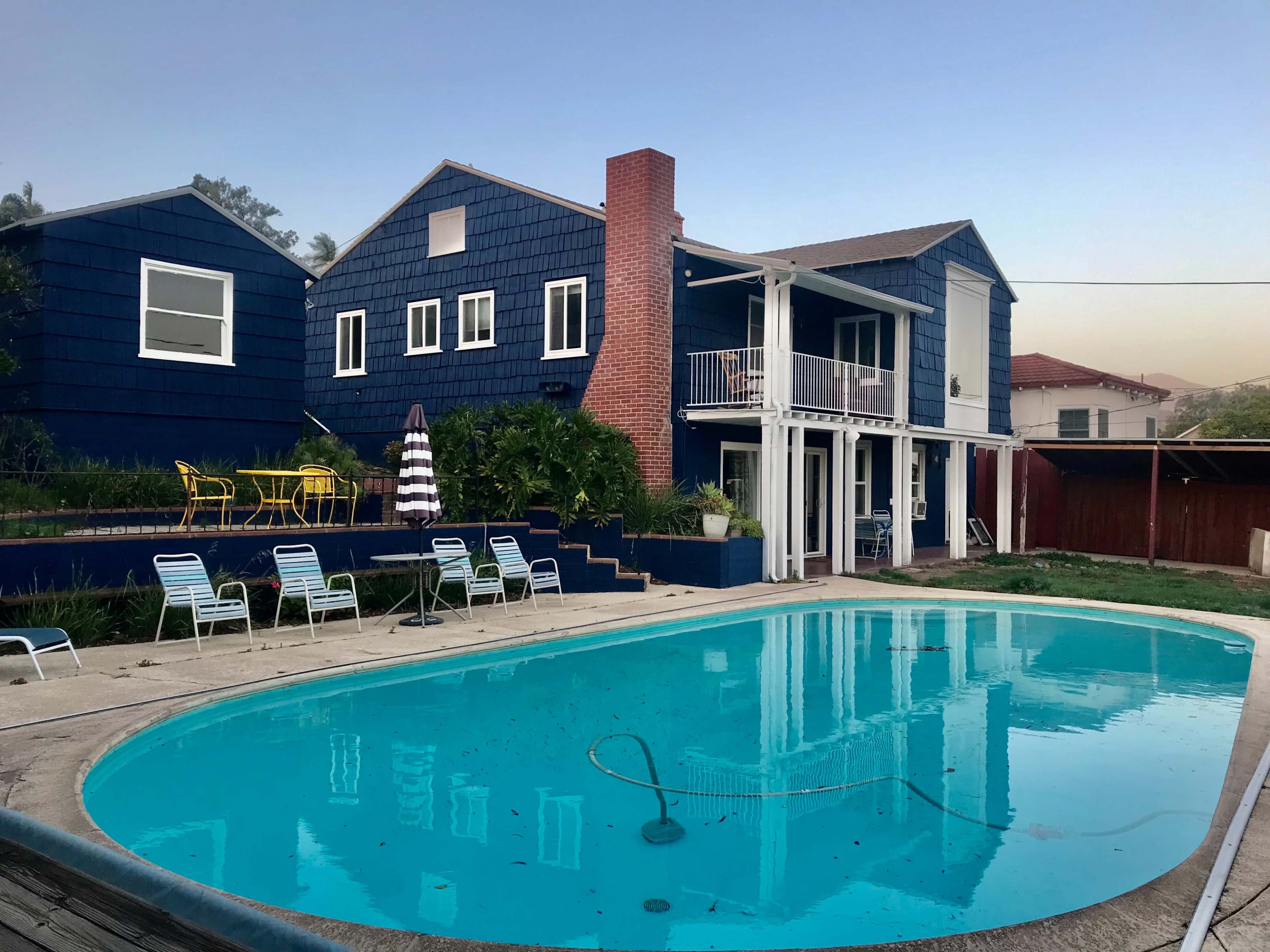 A rectangular pool reflects a blue two-story house with a chimney and outdoor seating.