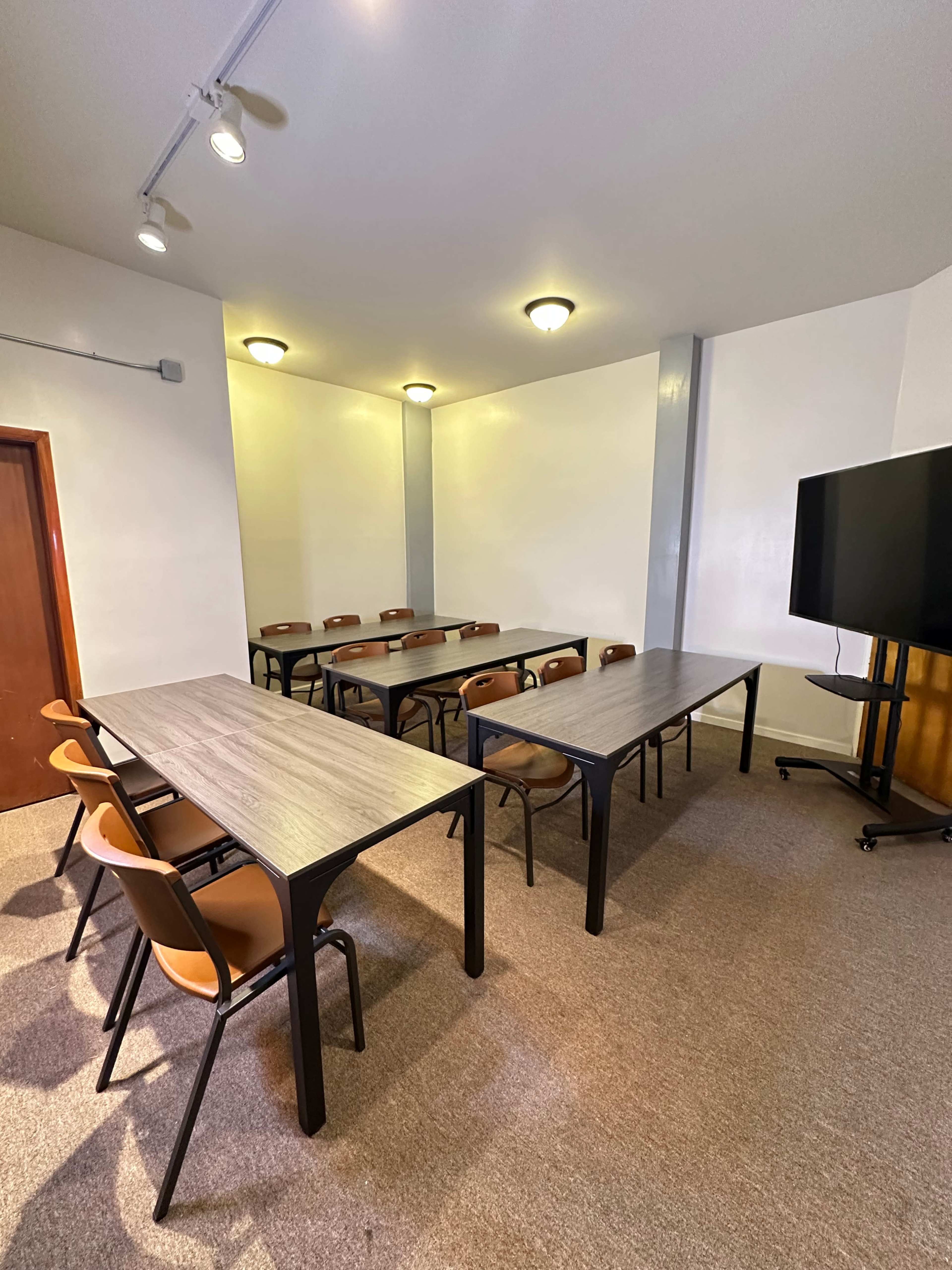 A conference room is arranged with several tables and chairs, and a large screen is positioned at one end.