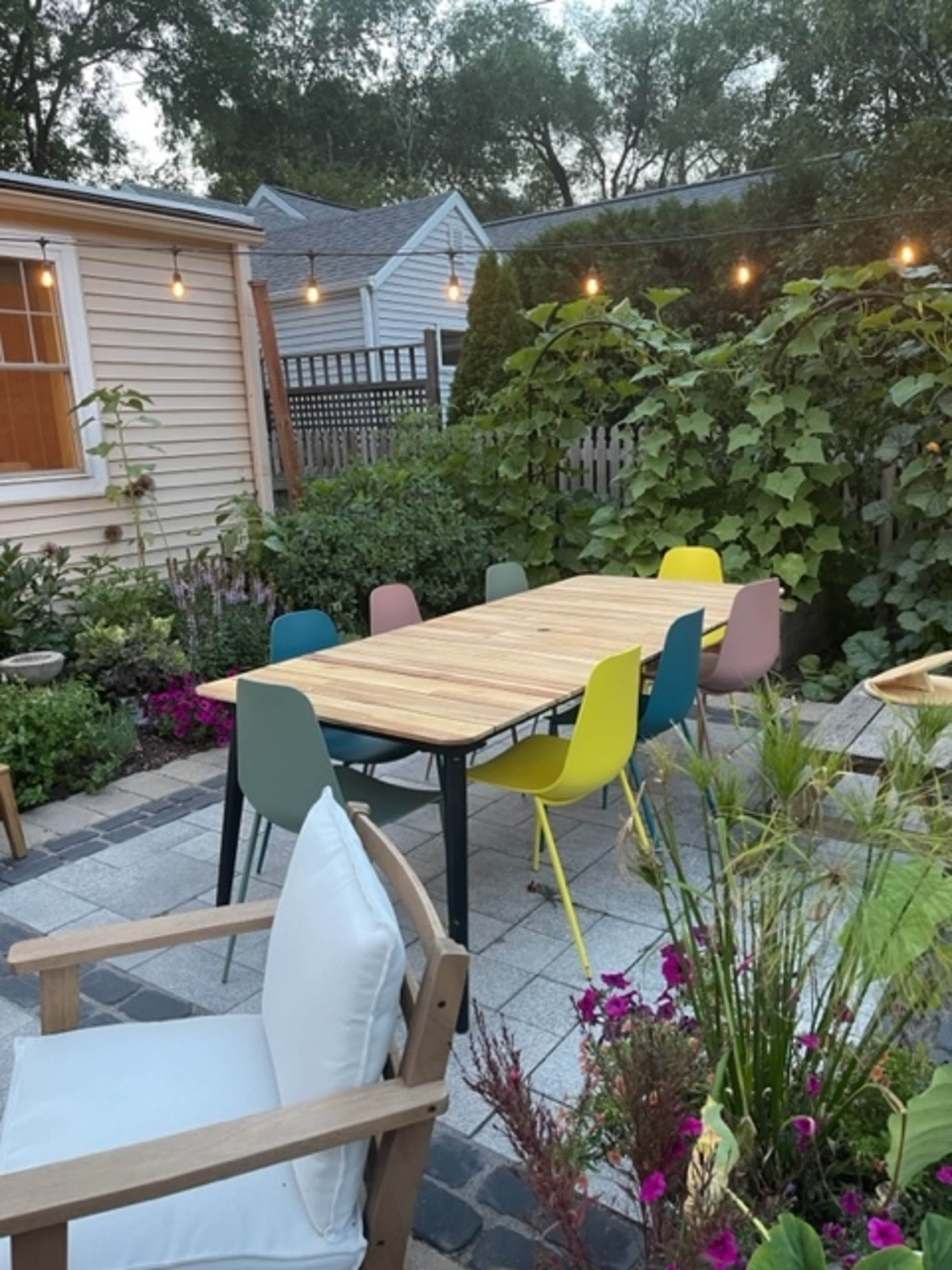 A wooden dining table surrounded by colorful chairs is set in a garden area with plants and flowers.