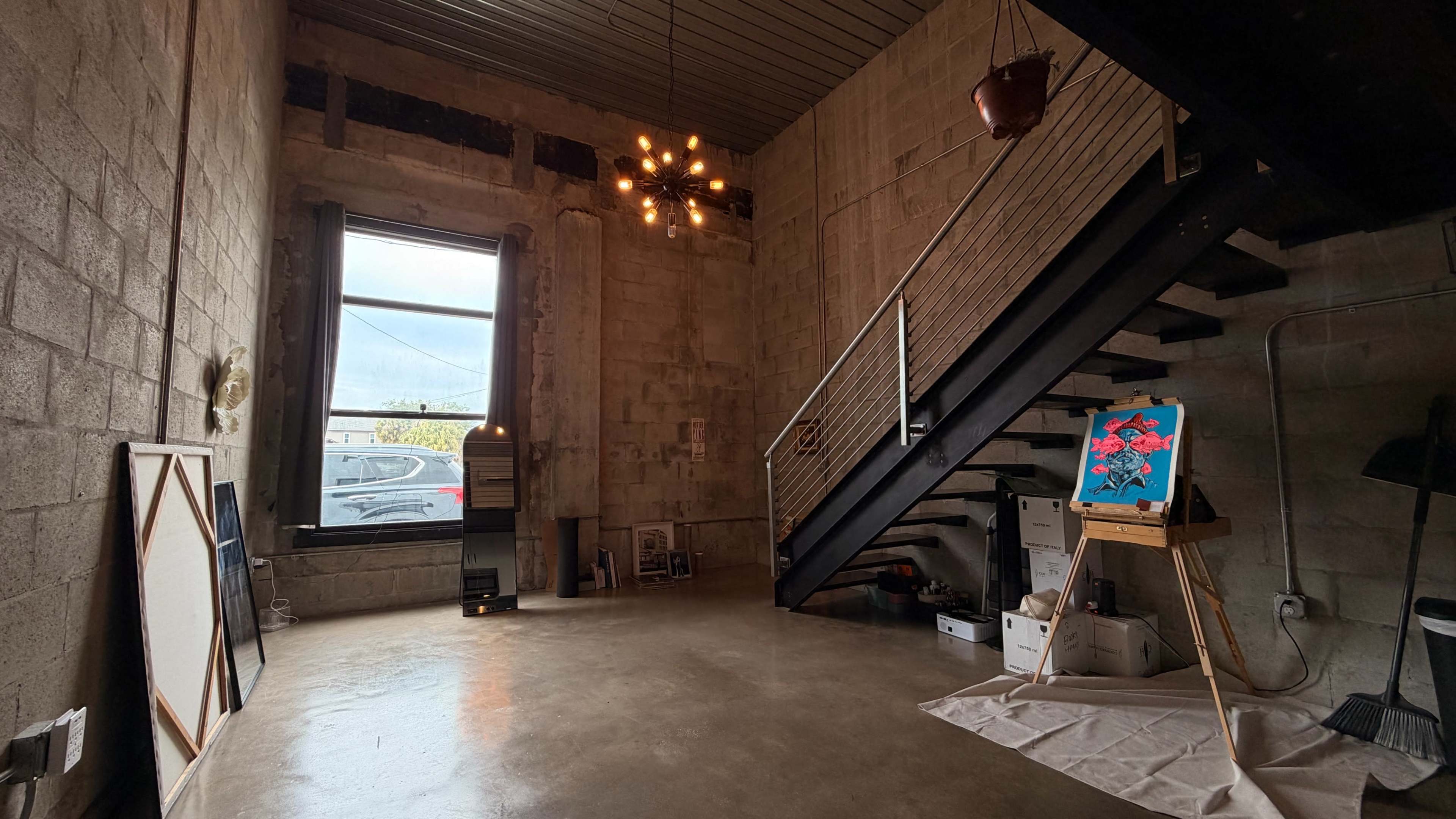 The image shows an industrial-style interior space with a staircase, art on an easel, and large windows providing natural light.