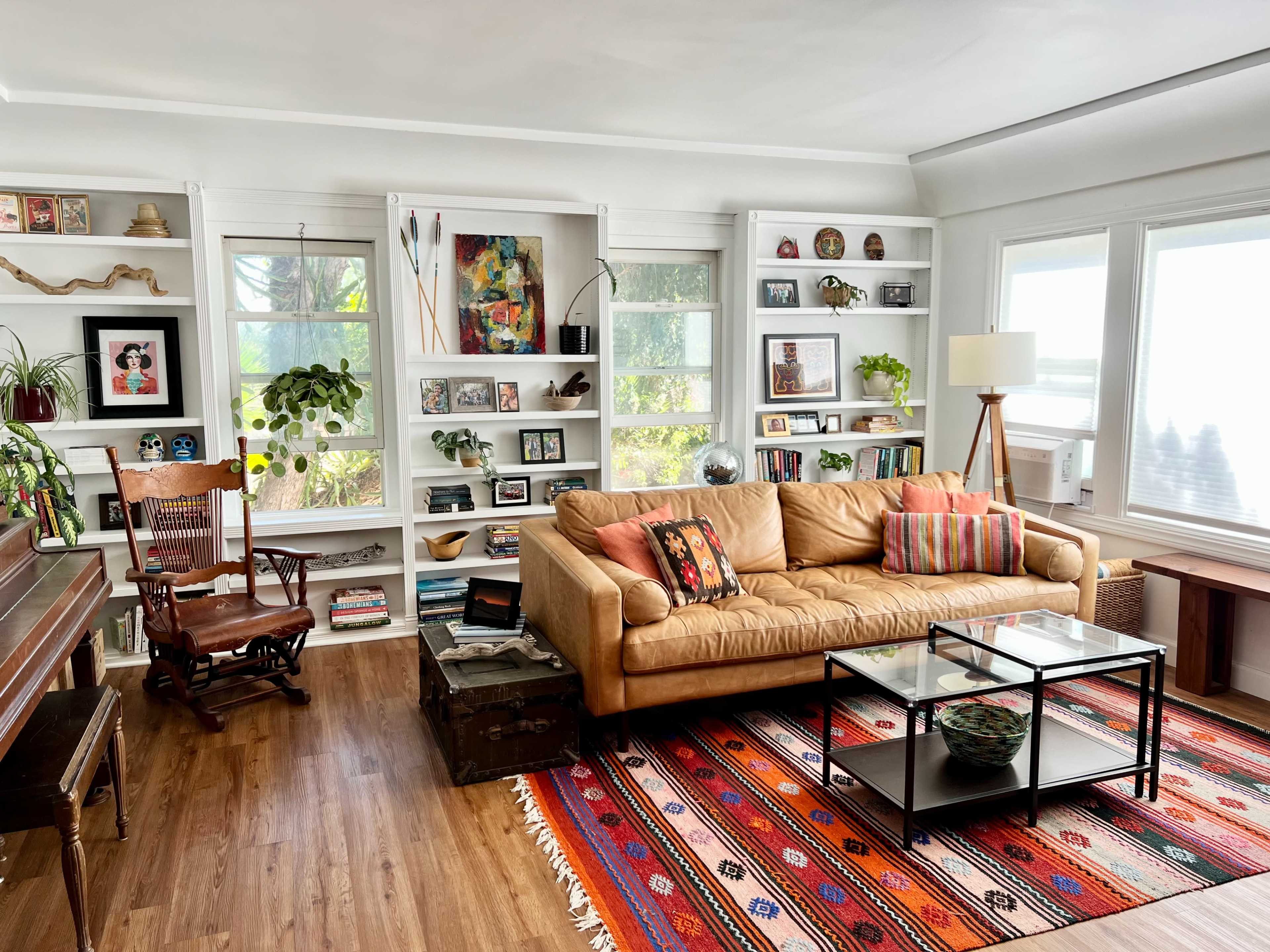 The living room features a tan leather sofa, a glass coffee table, bookshelves lined with books and decorative items, a rocking chair, and large windows allowing natural light.