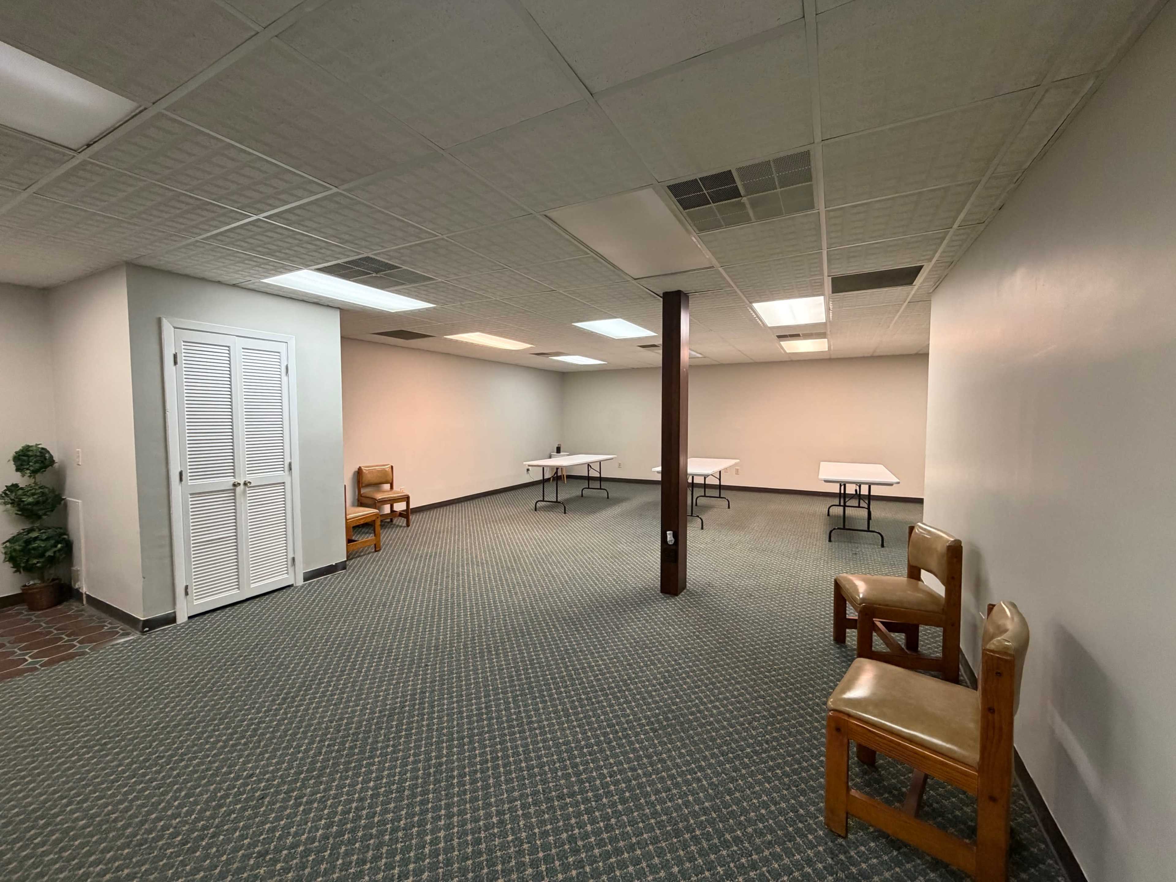 The image shows an empty room with two chairs against one wall, two folding tables in the center, and a pair of closed double doors.
