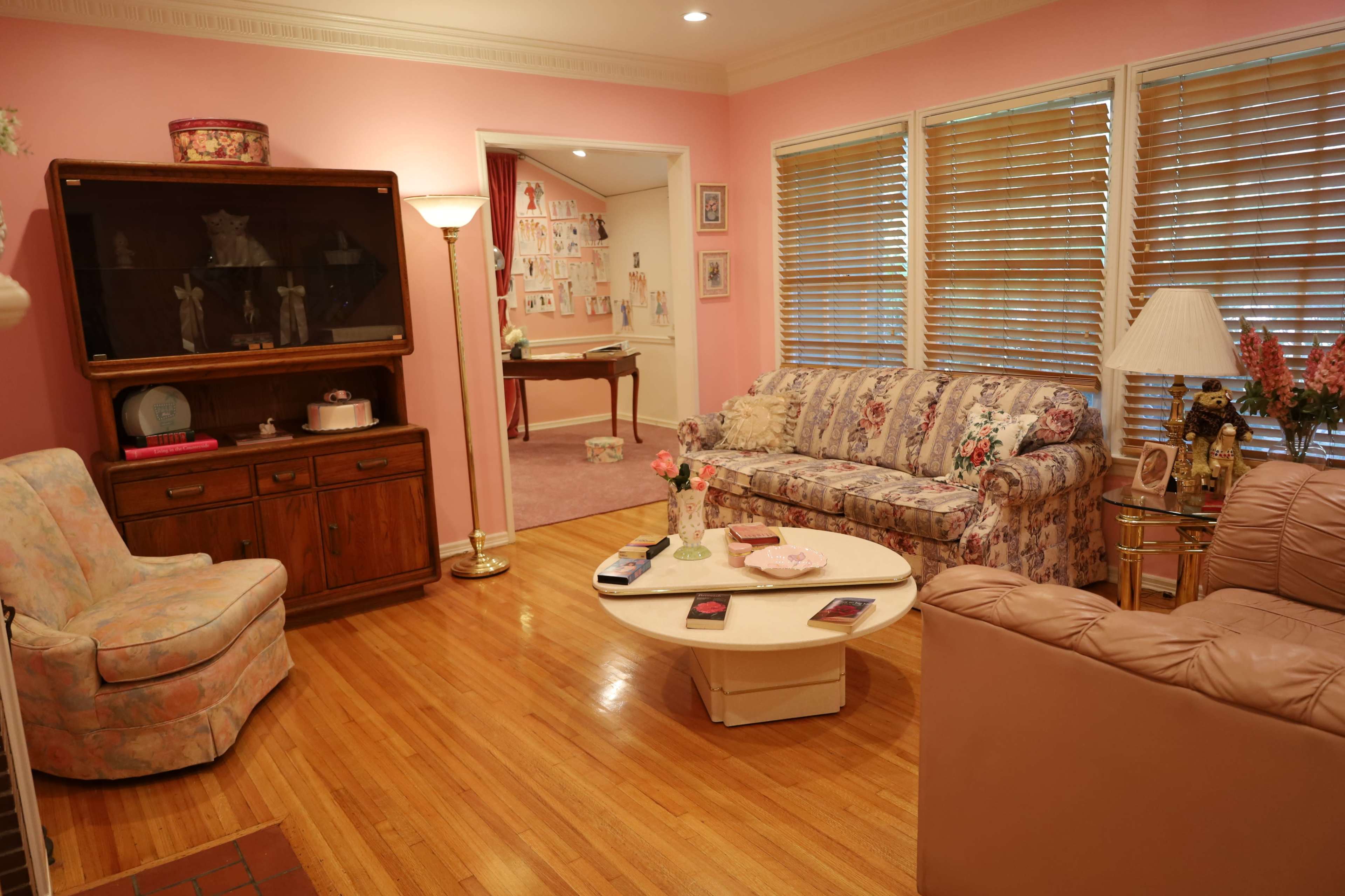 The living room features floral-patterned sofas, a wooden cabinet, a round coffee table, and pink walls with wooden blinds.