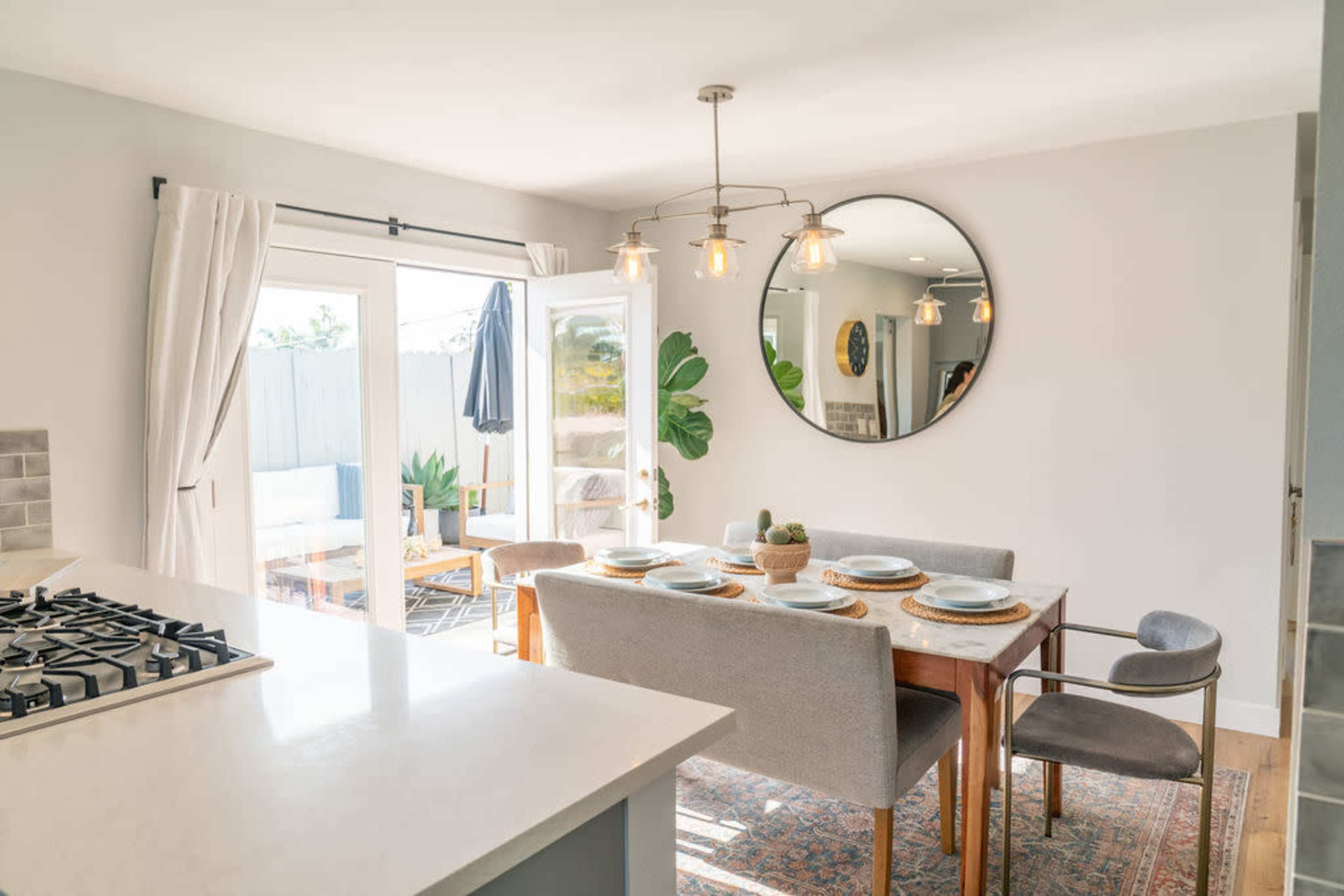 A dining area features a wooden table set with plates, surrounded by chairs, with large windows and a mirror reflecting the light.