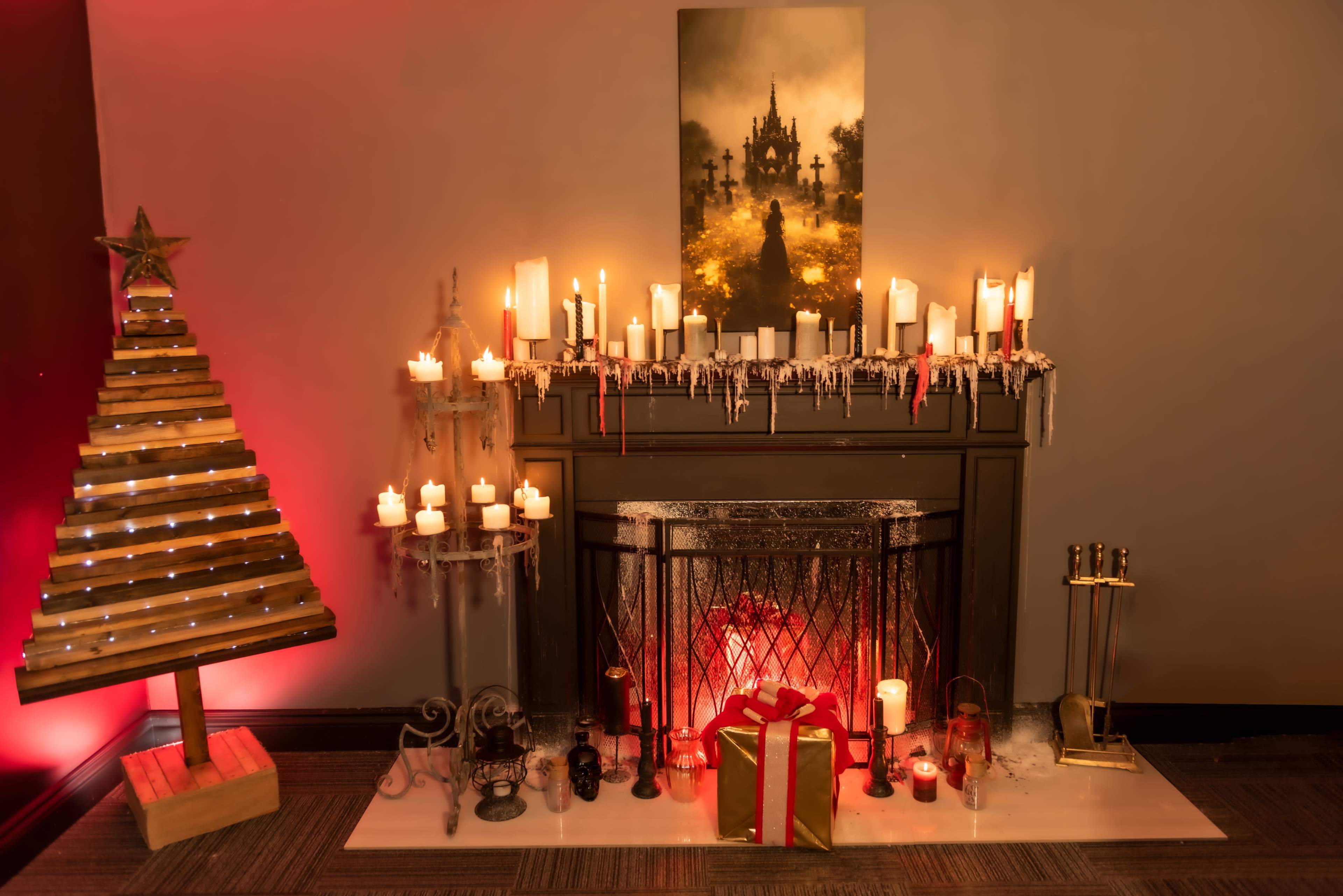 A decorated fireplace features candles, a gift, and a wooden Christmas tree, all set against a backdrop of a misty landscape print.