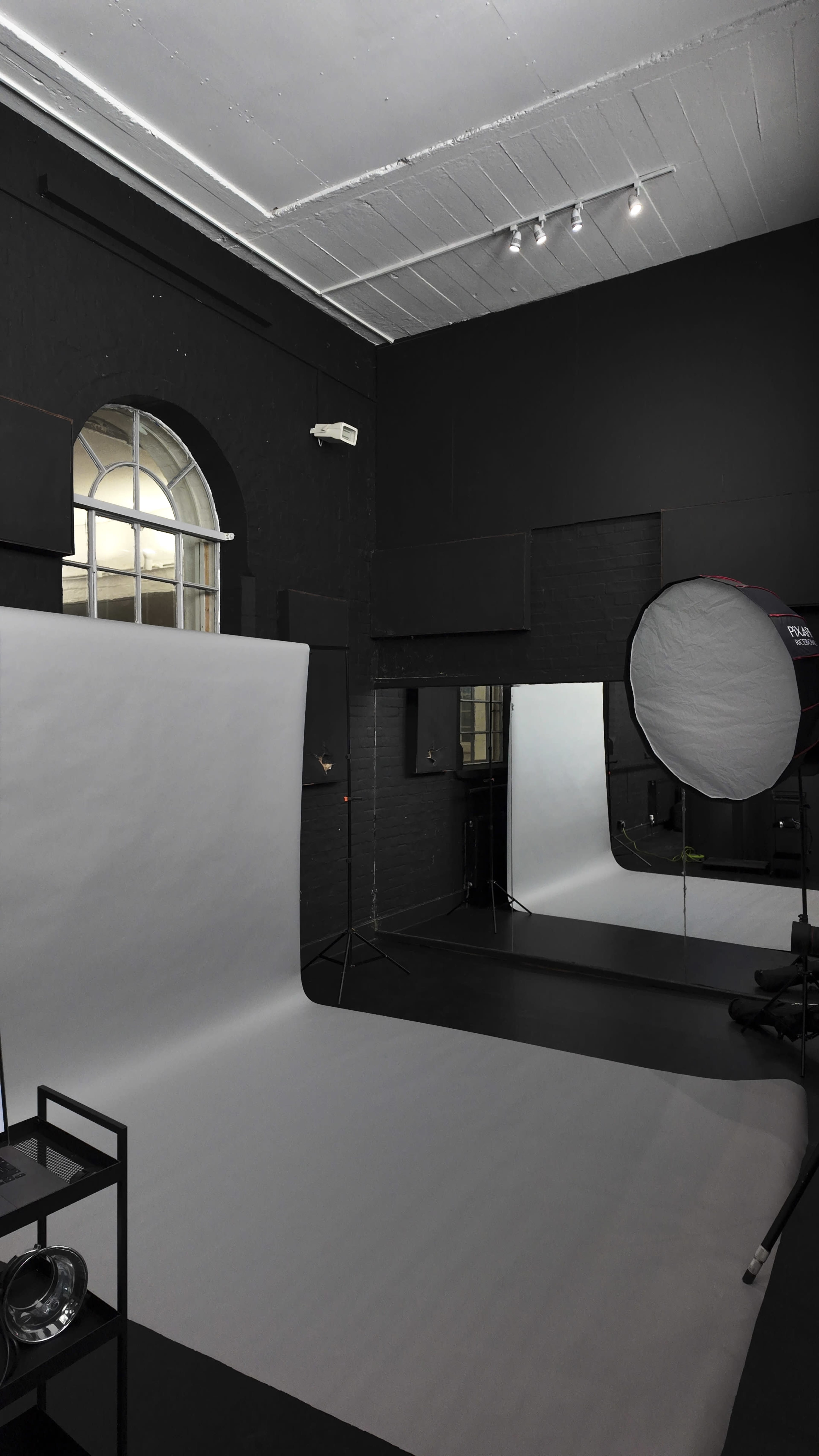 The image shows a photography studio with grey seamless backdrop paper, a round reflector, and minimal furnishings against black walls.