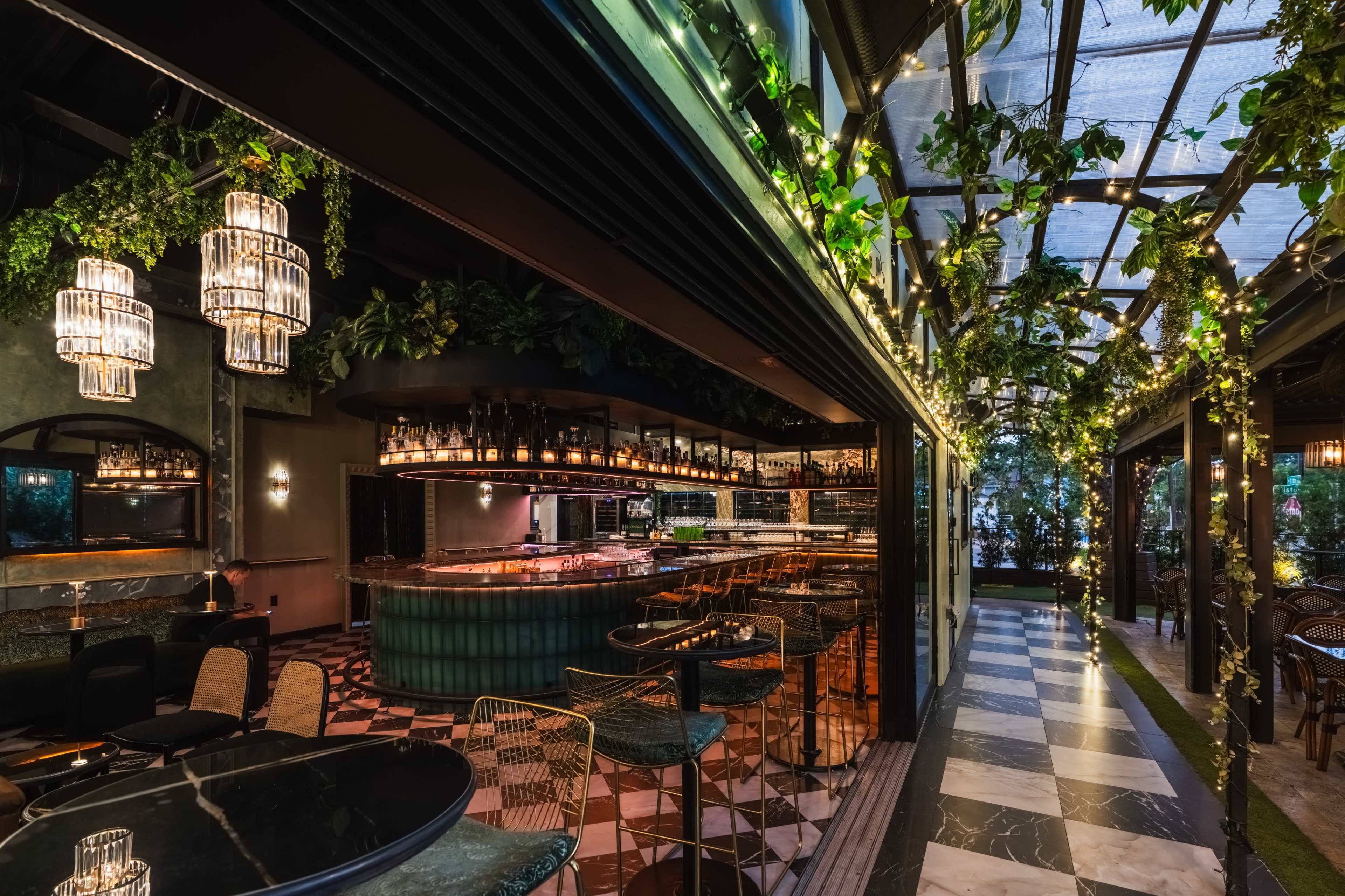 The image shows a modern restaurant interior featuring a bar with an illuminated counter, surrounded by greenery and decorative lights.