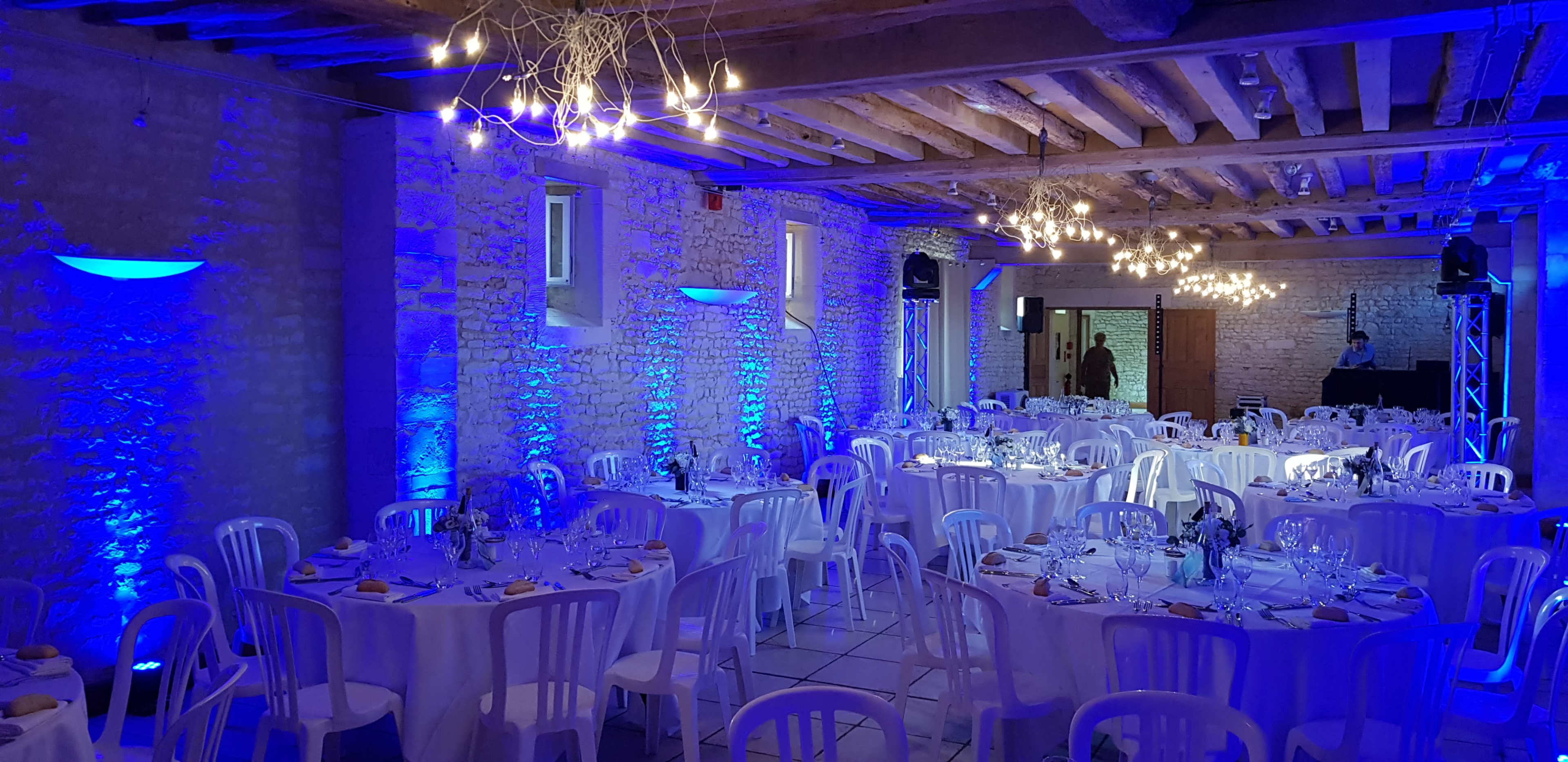 The image shows a banquet hall set with round tables covered in white tablecloths, illuminated by blue lighting from the walls and ceiling fixtures.