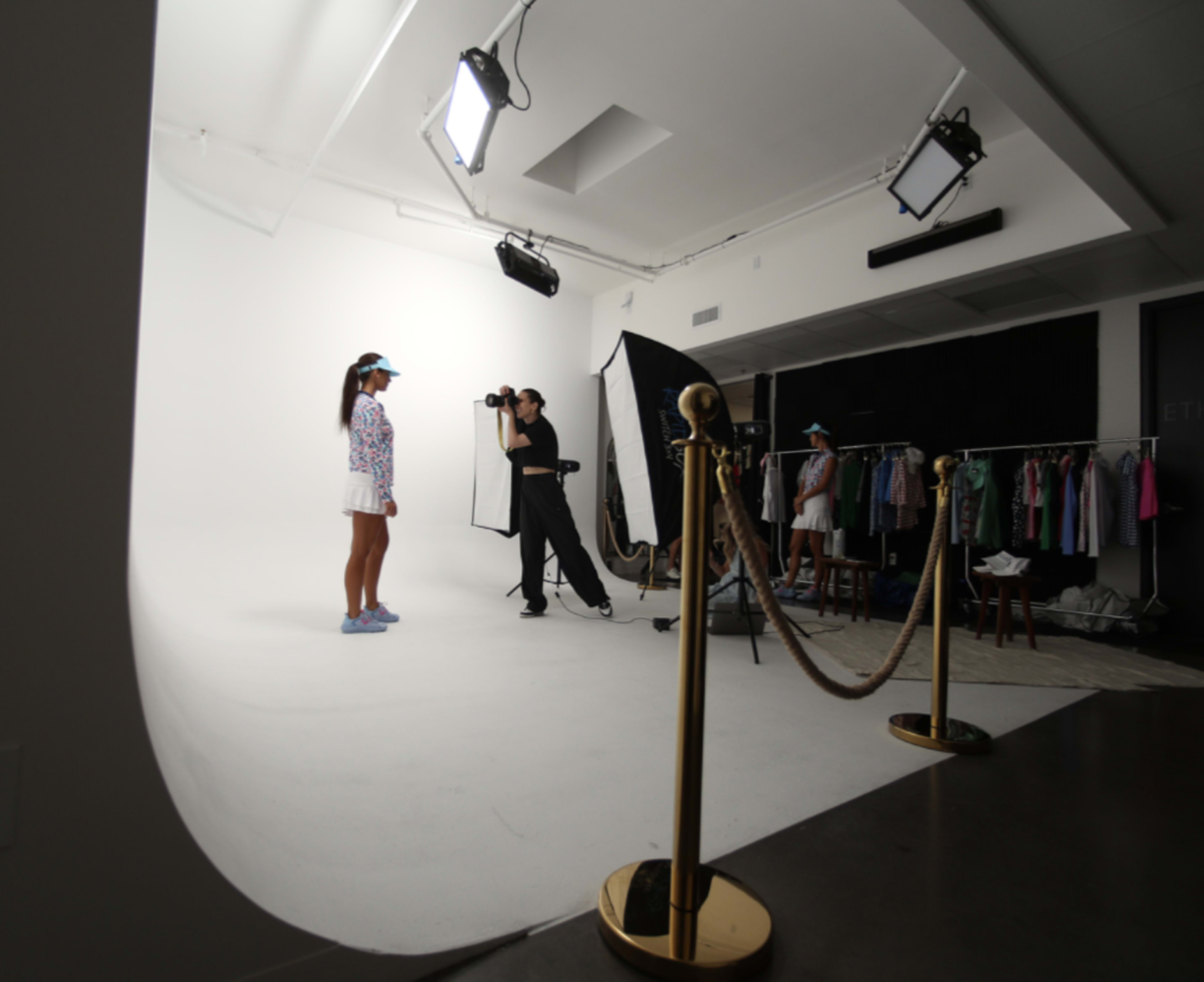 A model stands in a well-lit studio while a photographer takes pictures, with clothing racks and a rope barrier in the background.