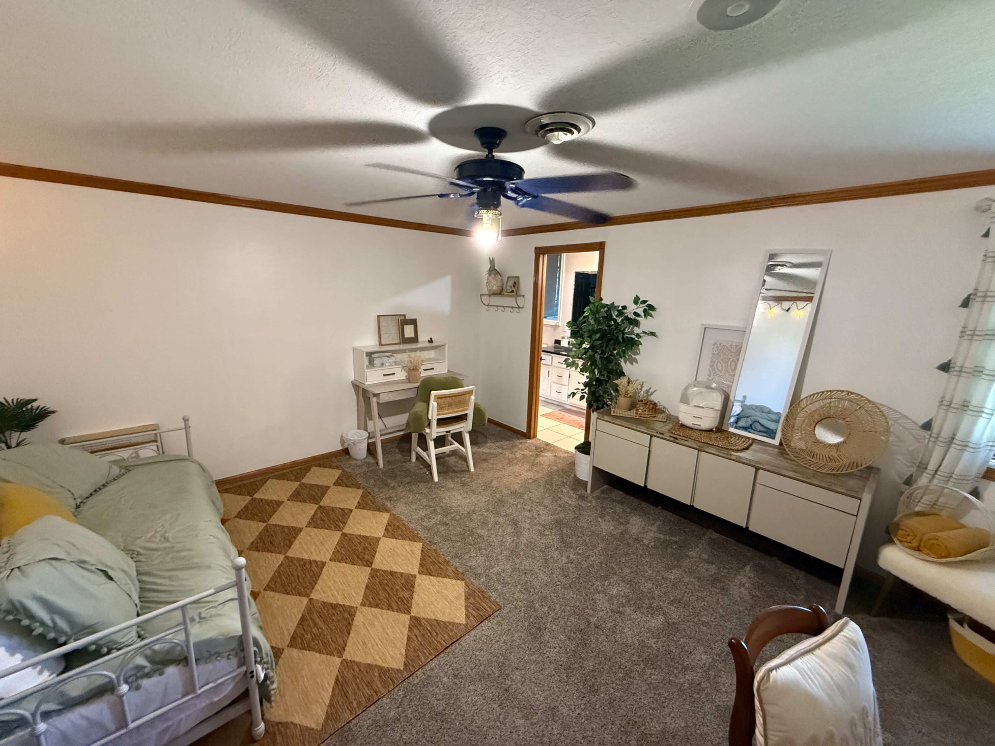 The image shows a softly lit bedroom with a bed, a desk and chair, a dresser, and a decorative plant in the corner.