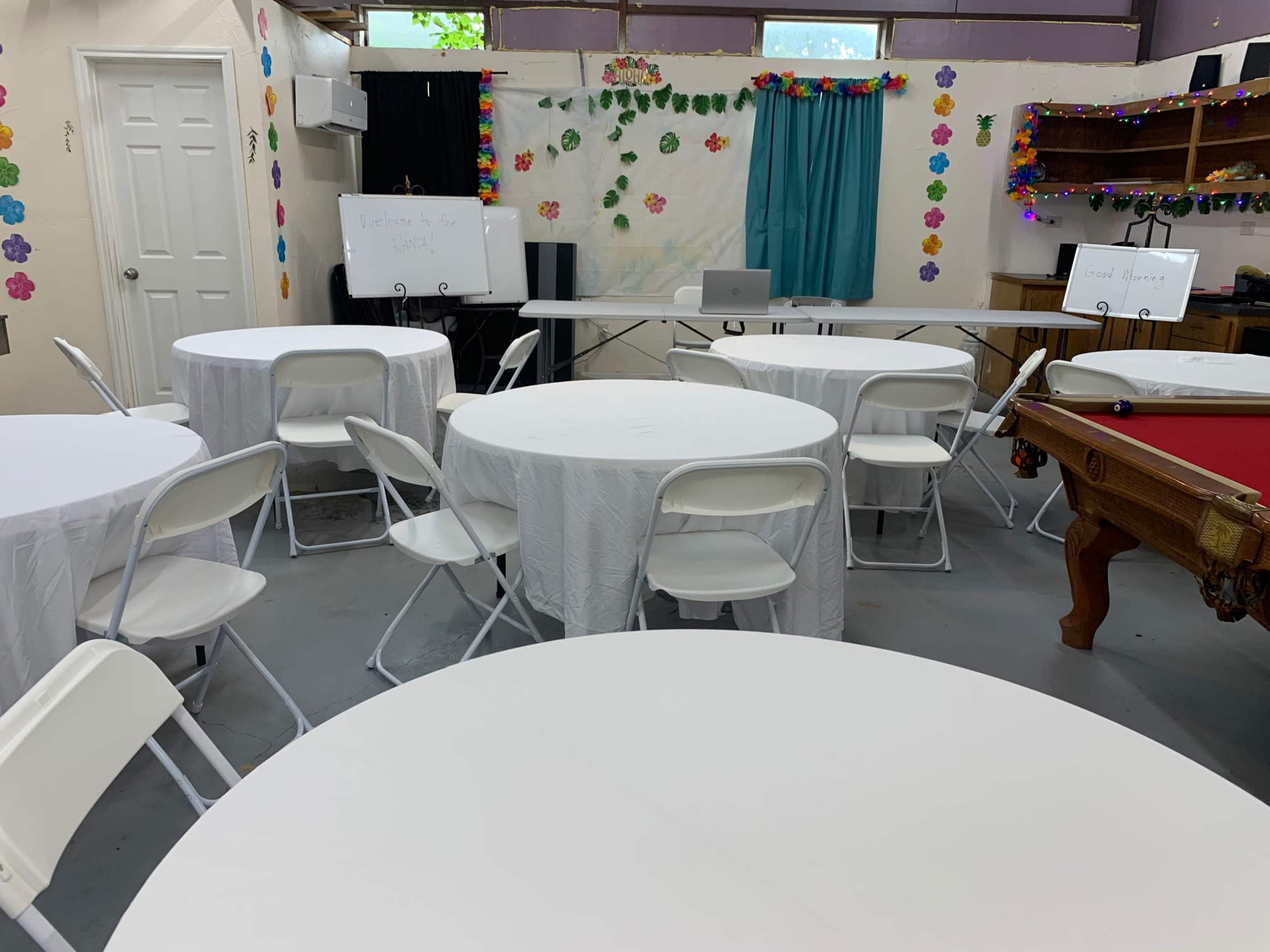 The image shows a room set up with several round tables covered in white tablecloths, accompanied by folding chairs, and featuring a projector screen and a chalkboard at the front.