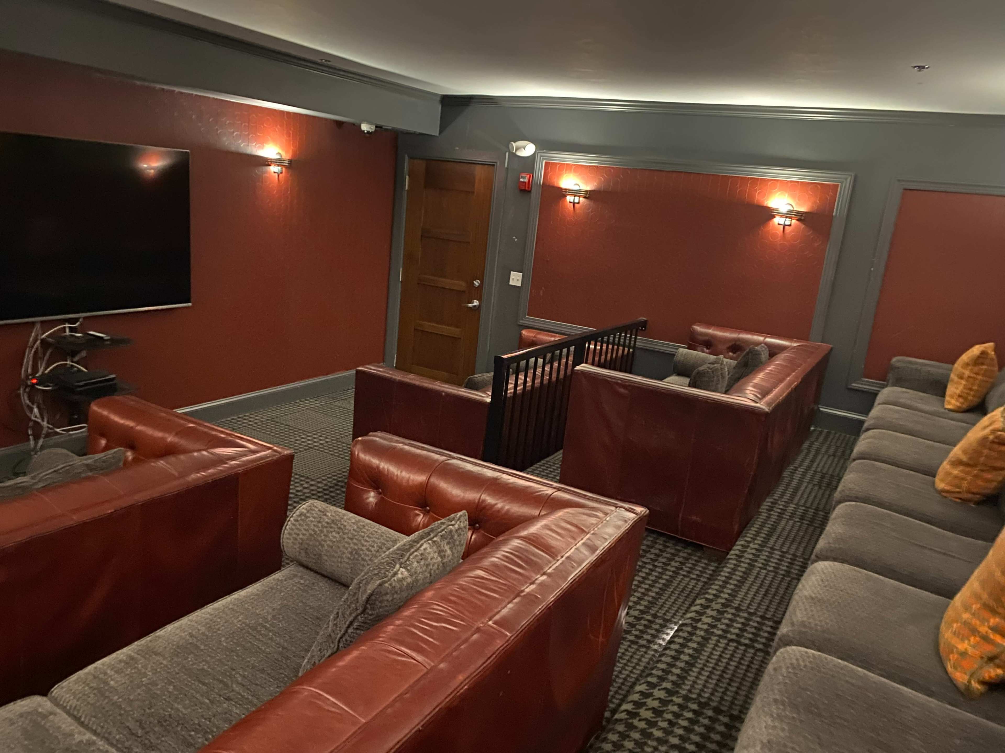 A cozy home theater room with red walls, leather sofas arranged in two sections, and a large wall-mounted TV.