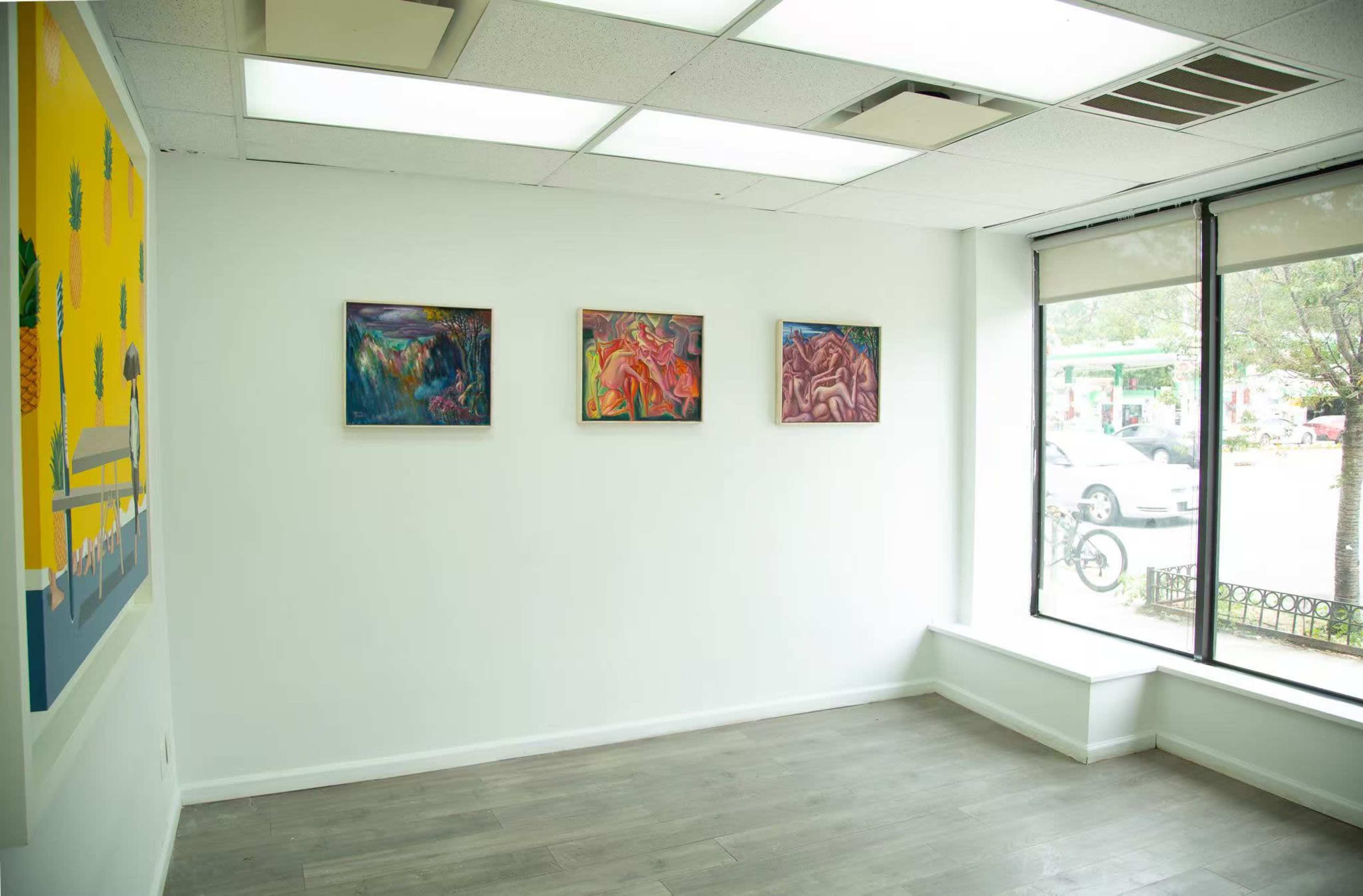 The image shows an empty gallery space with three pieces of artwork mounted on a white wall, next to a large window overlooking an outdoor area.