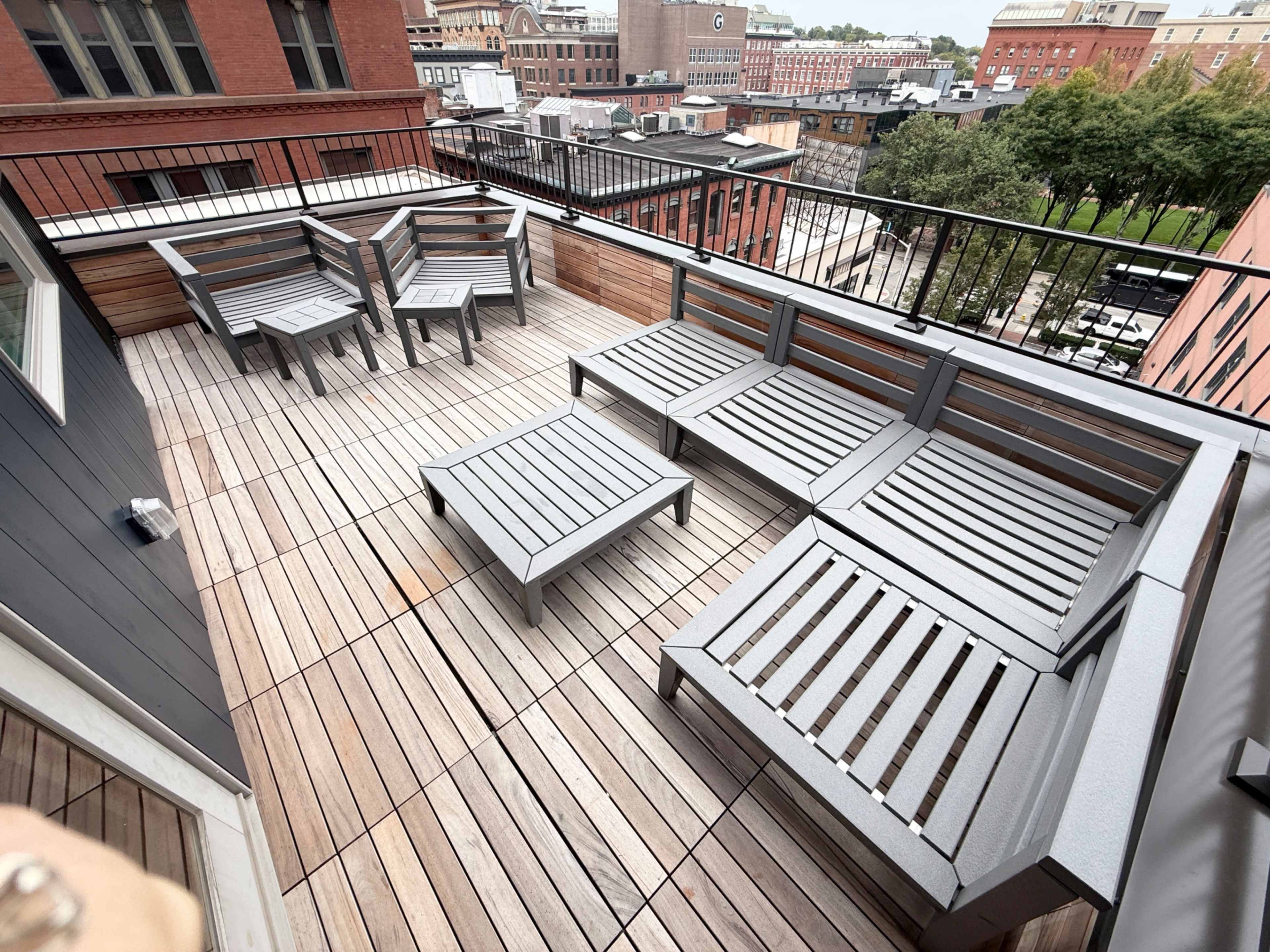 The image shows a rooftop terrace with wooden decking, featuring several metal-framed lounge chairs and a small table.