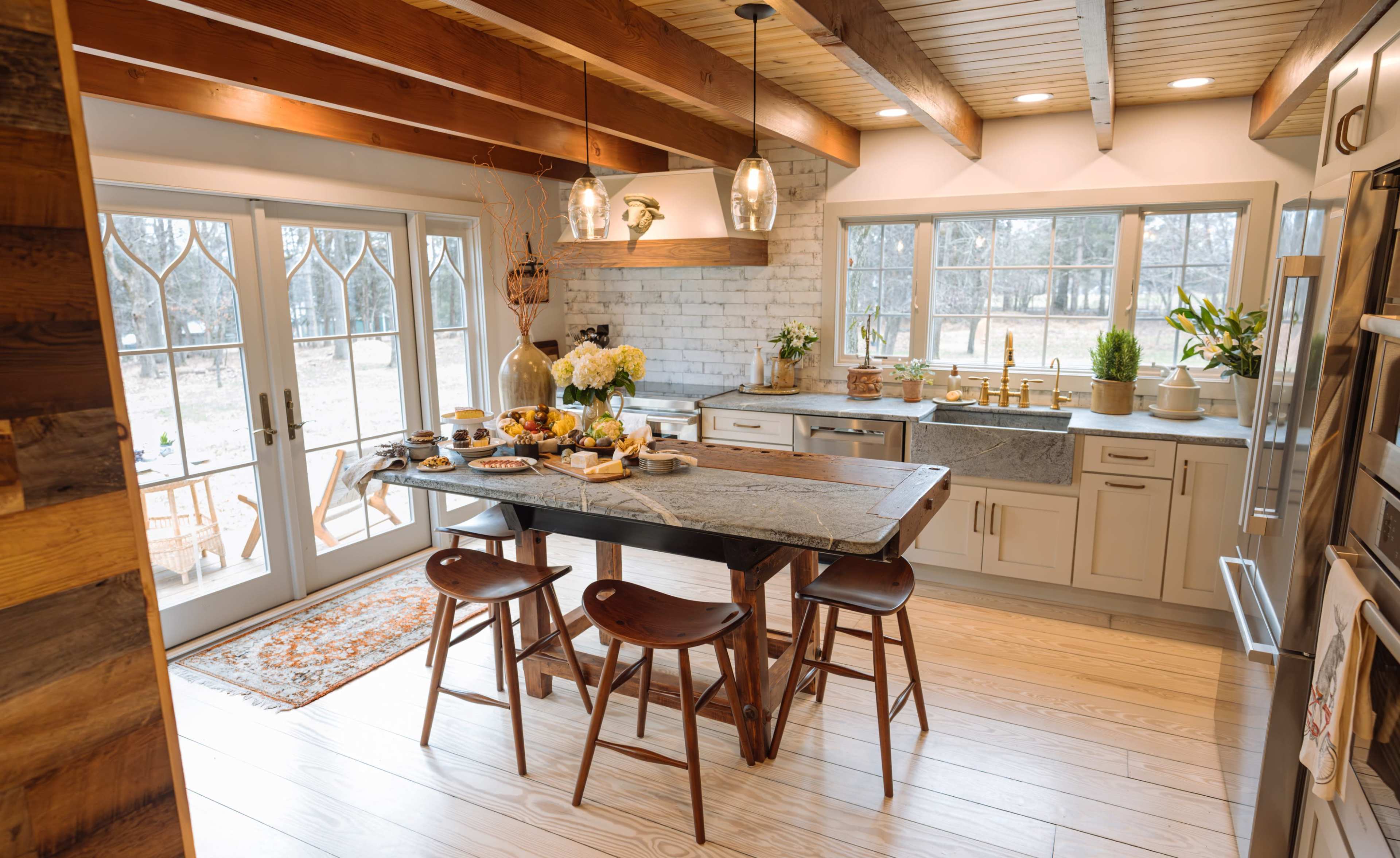 A modern kitchen features a large wooden dining table, white cabinetry, and large windows overlooking a patio area.