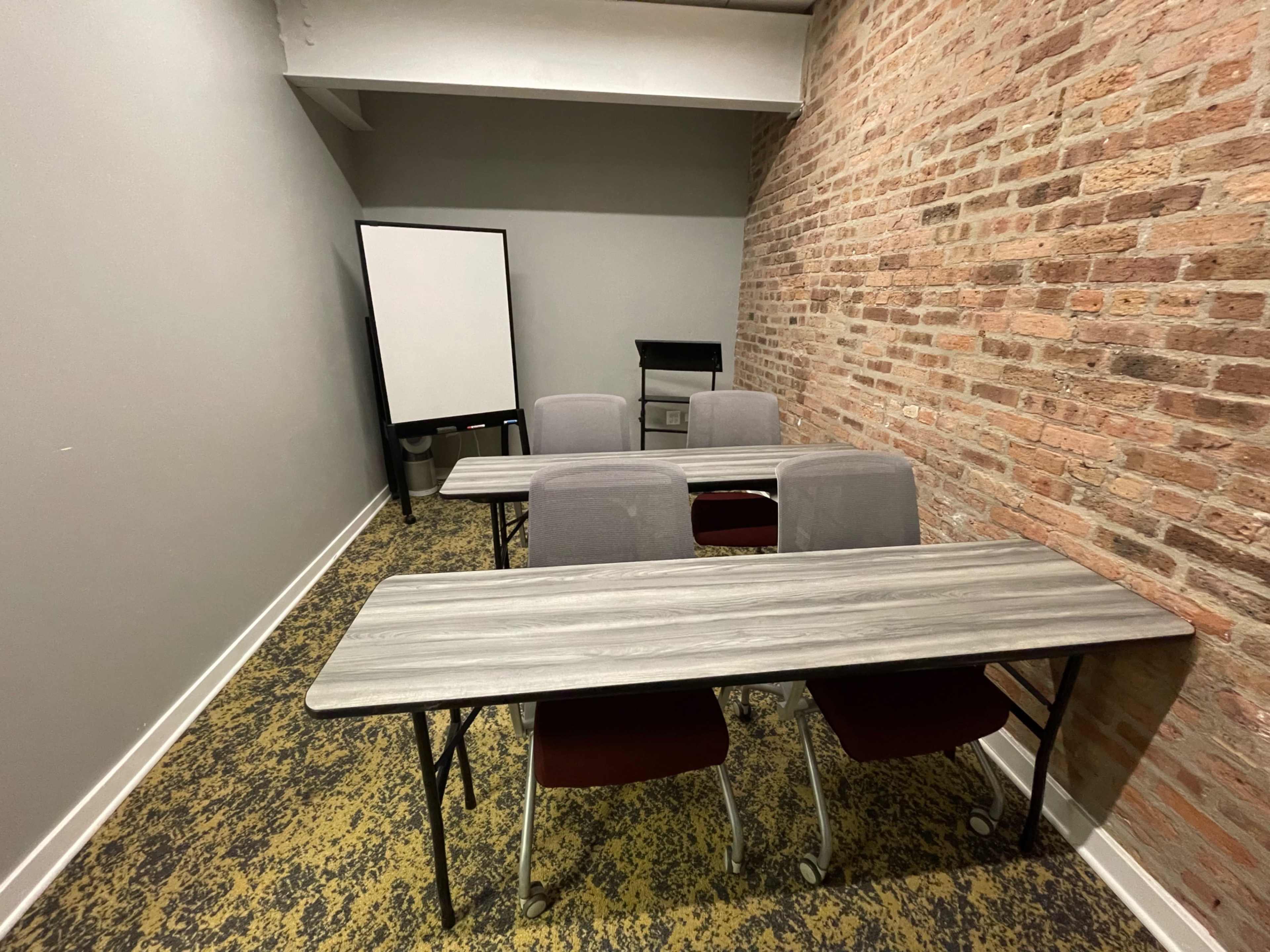 The image shows a small meeting room with four gray chairs around two rectangular tables, a whiteboard on an easel, and brick walls.