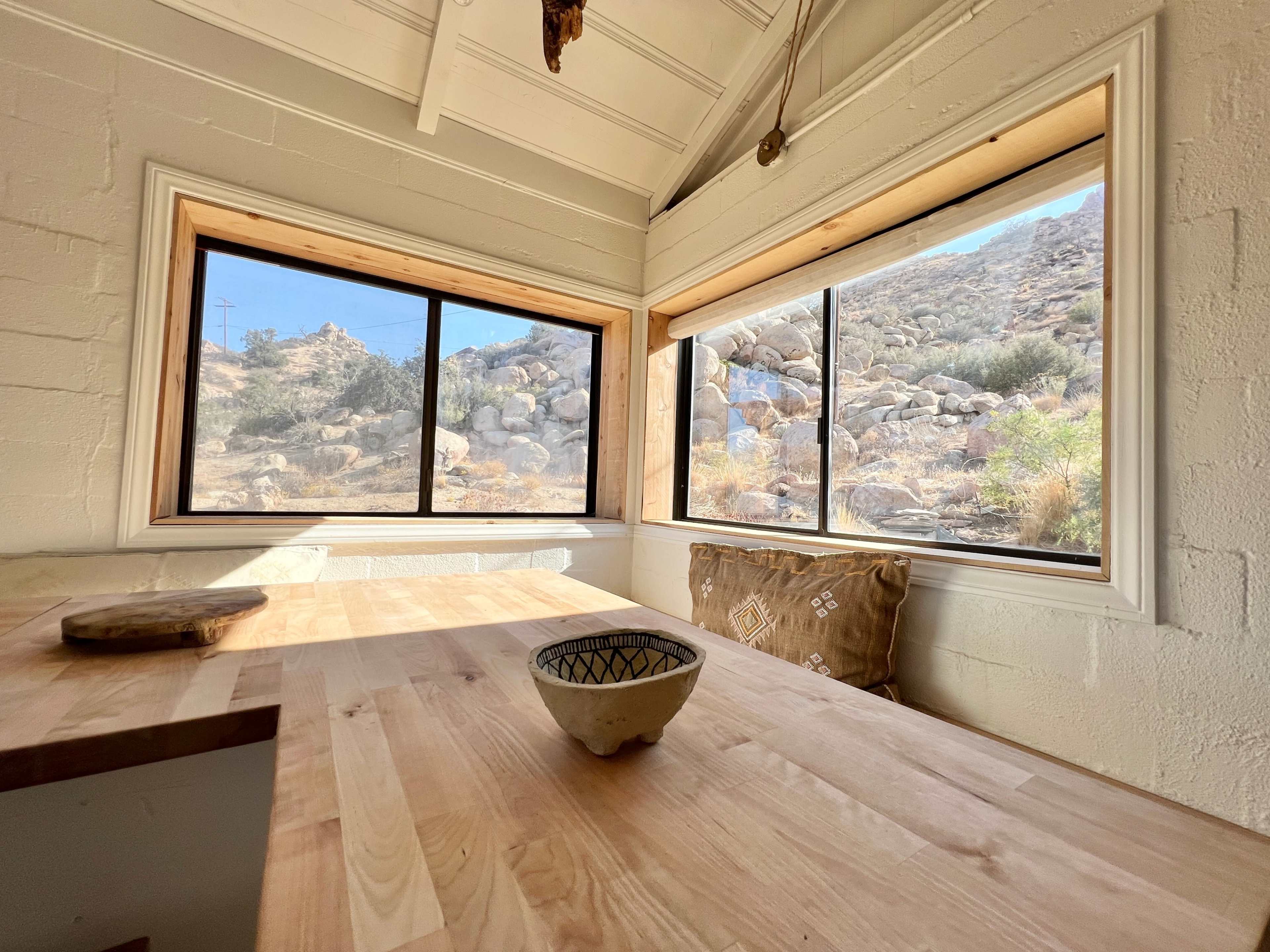 A wooden table with a ceramic bowl sits next to large windows that offer a view of a rocky hillside.