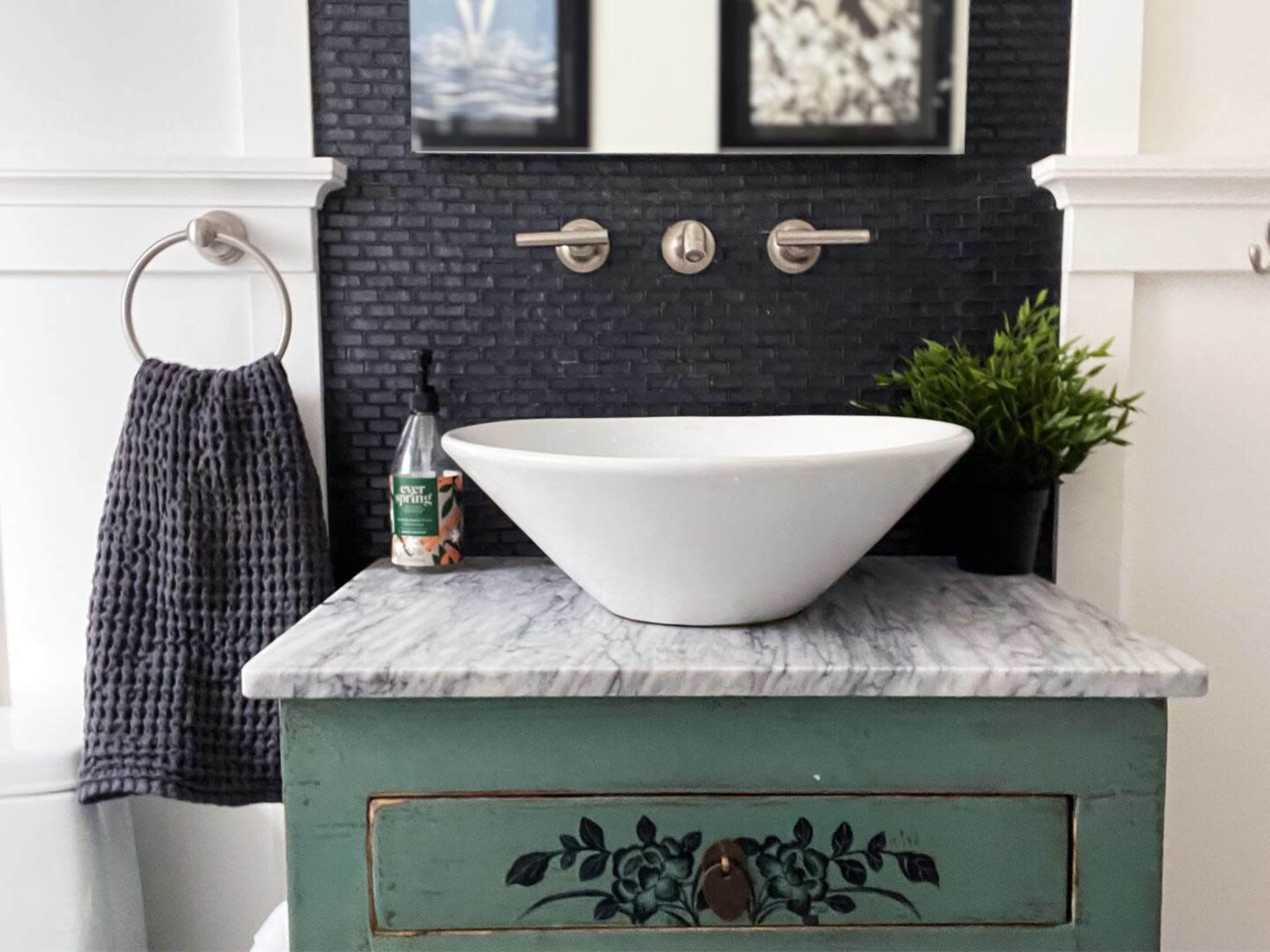 A modern bathroom vanity with a white vessel sink on a marble countertop, a green cabinet below, and wall-mounted faucets above.