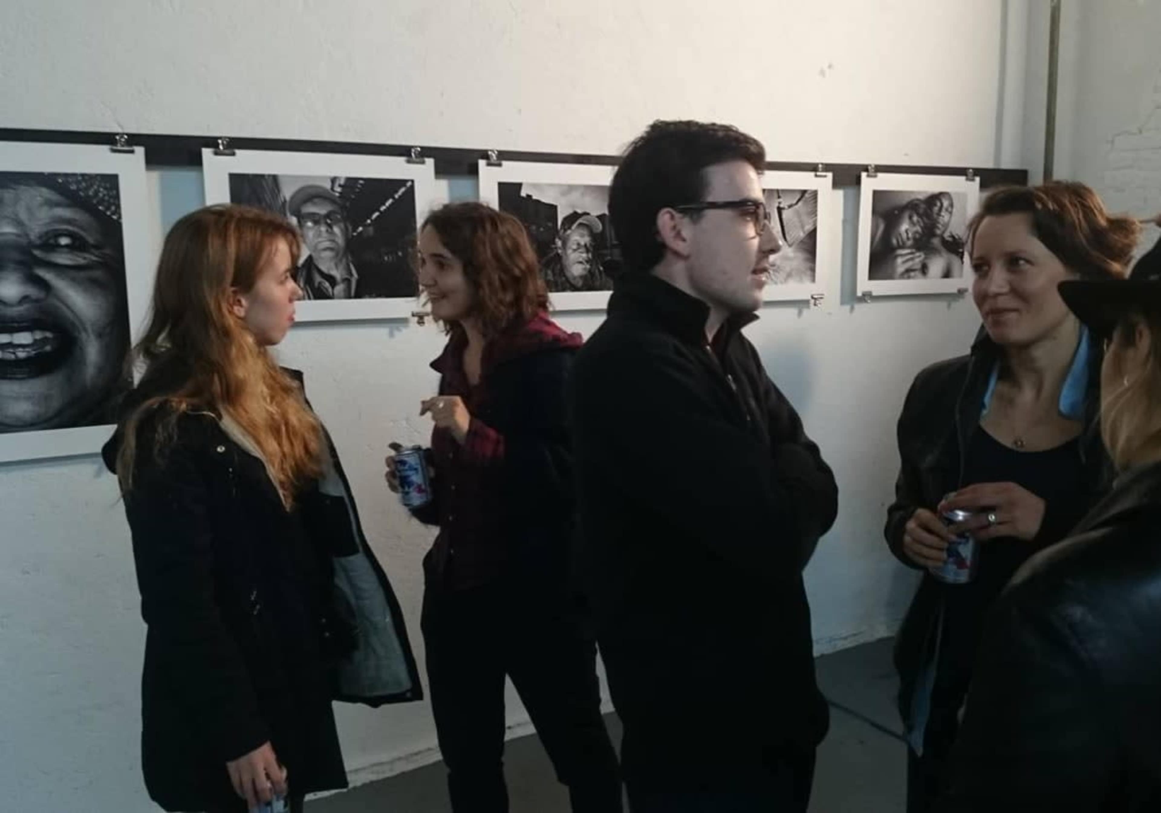 A group of four people converses in an art gallery featuring black-and-white photographs on the walls.
