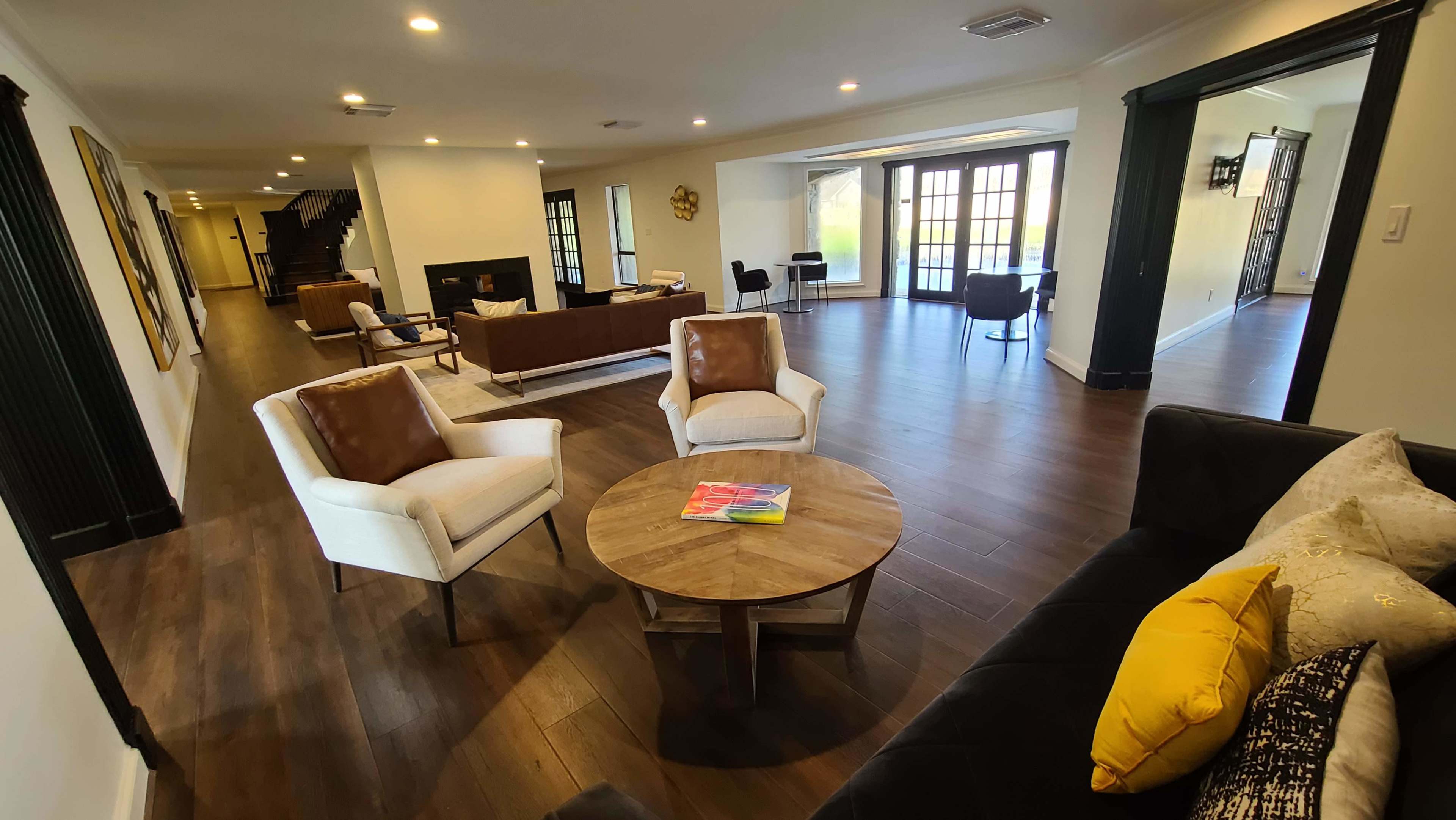 The image shows a spacious, modern living area with hardwood floors, featuring a round wooden coffee table, two armchairs, and a view of an adjacent room with couches and large windows.