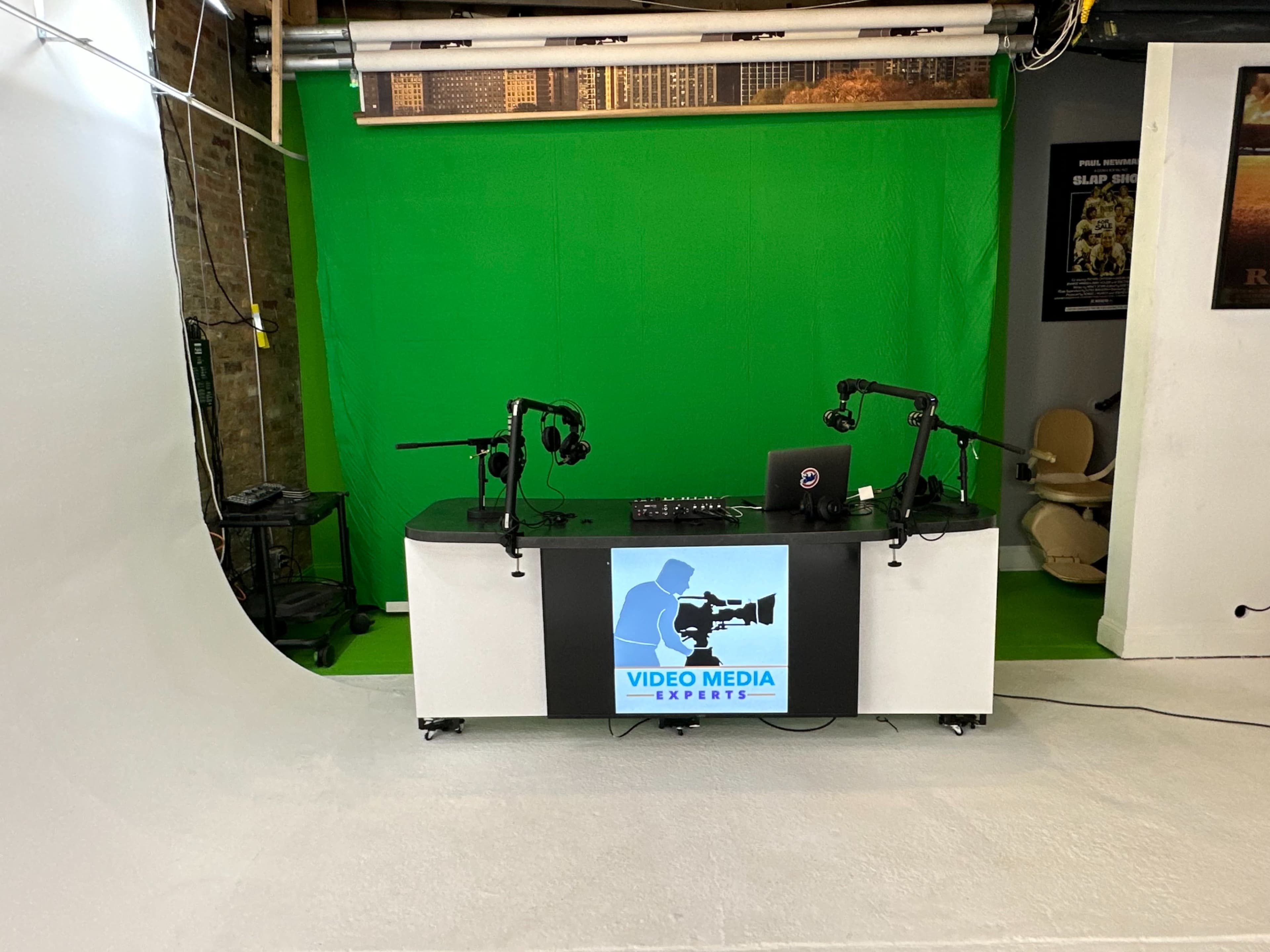 A video production studio features a green screen backdrop, a central black and white desk with a logo, and two microphone stands.