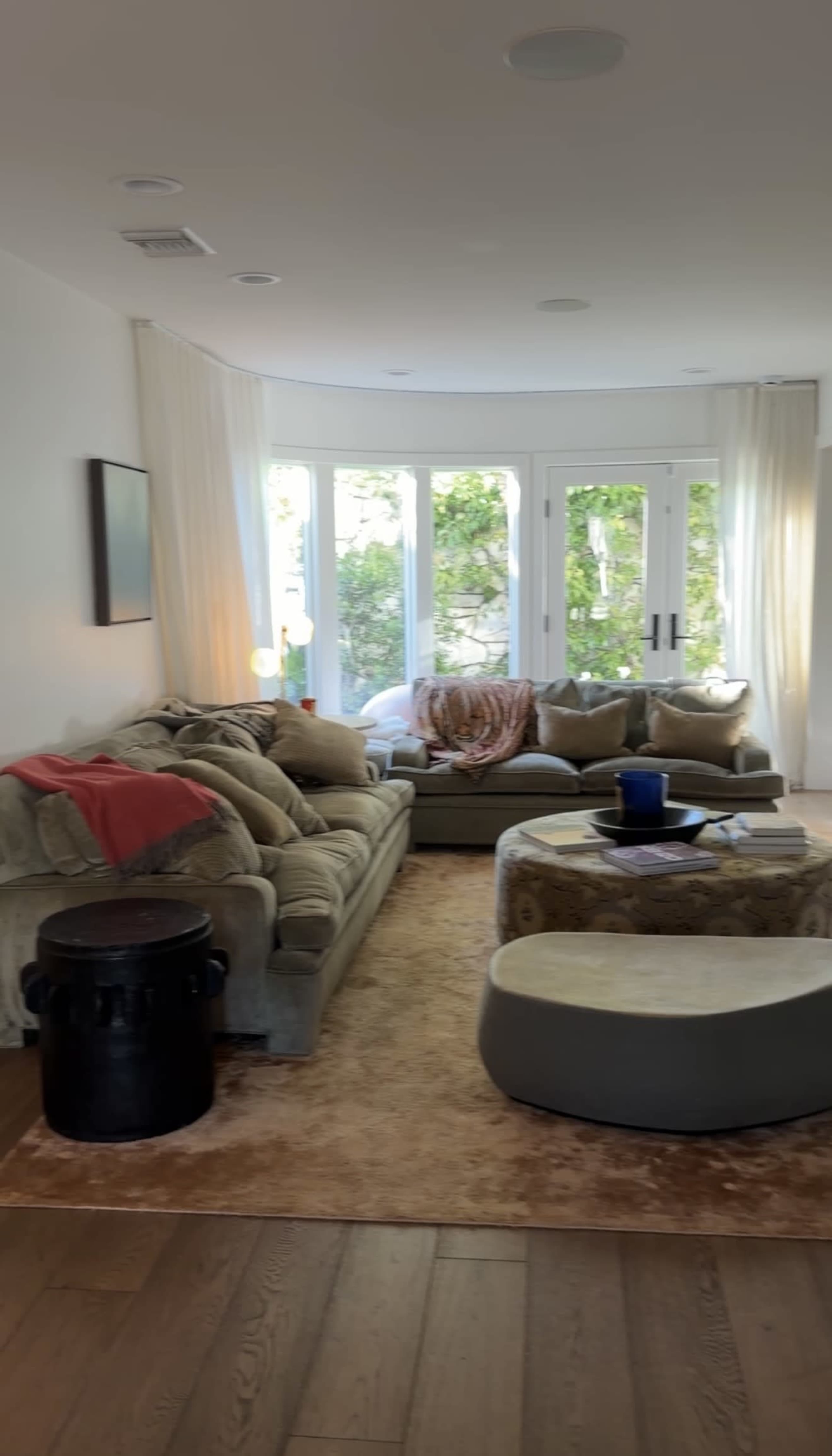 The image shows a living room with a sectional sofa, a round coffee table, and large windows letting in natural light.
