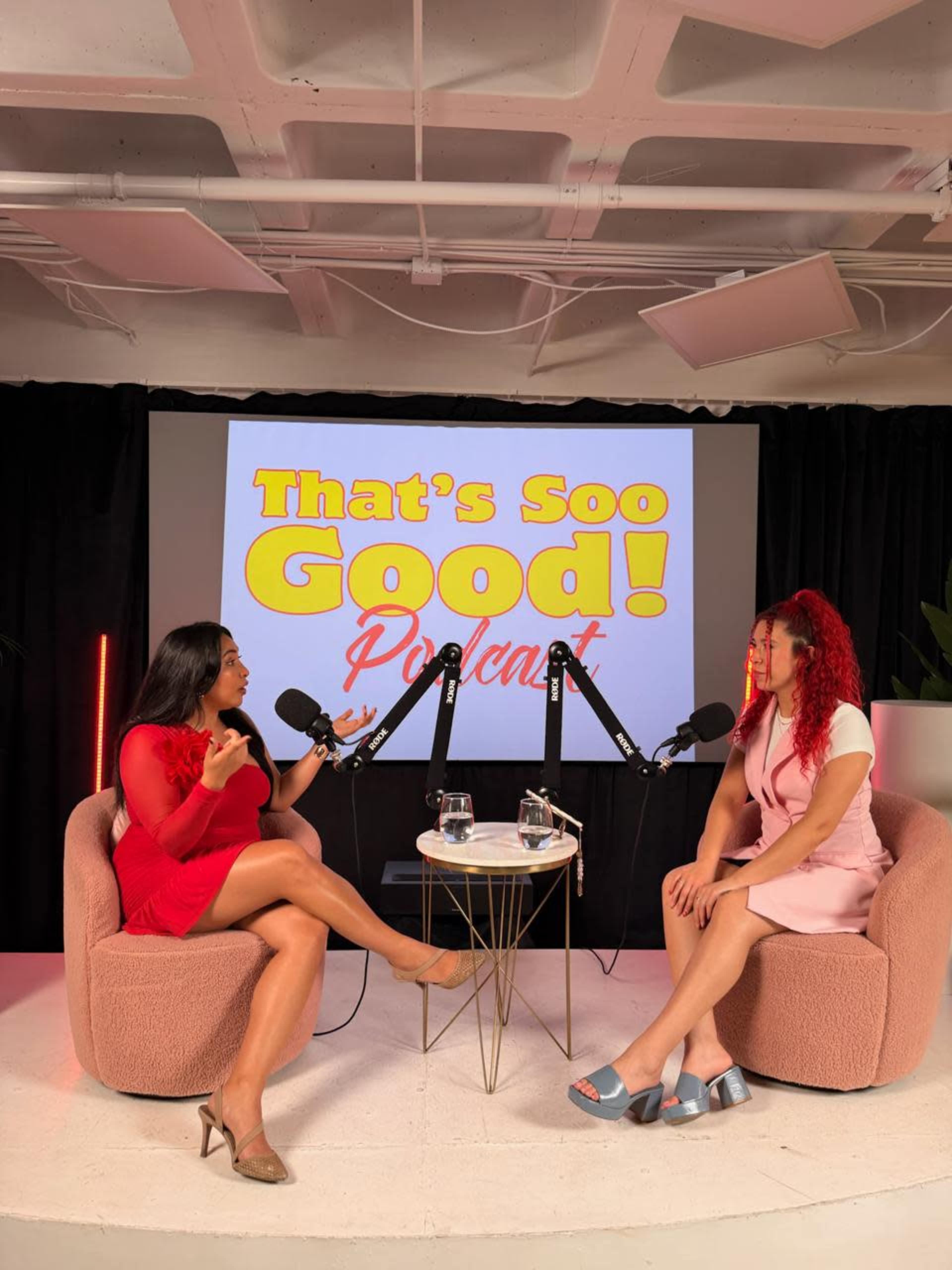 Two women are seated in a modern podcast studio, engaging in conversation while holding microphones, with a large screen displaying the title "That's Soo Good! Podcast" in the background.