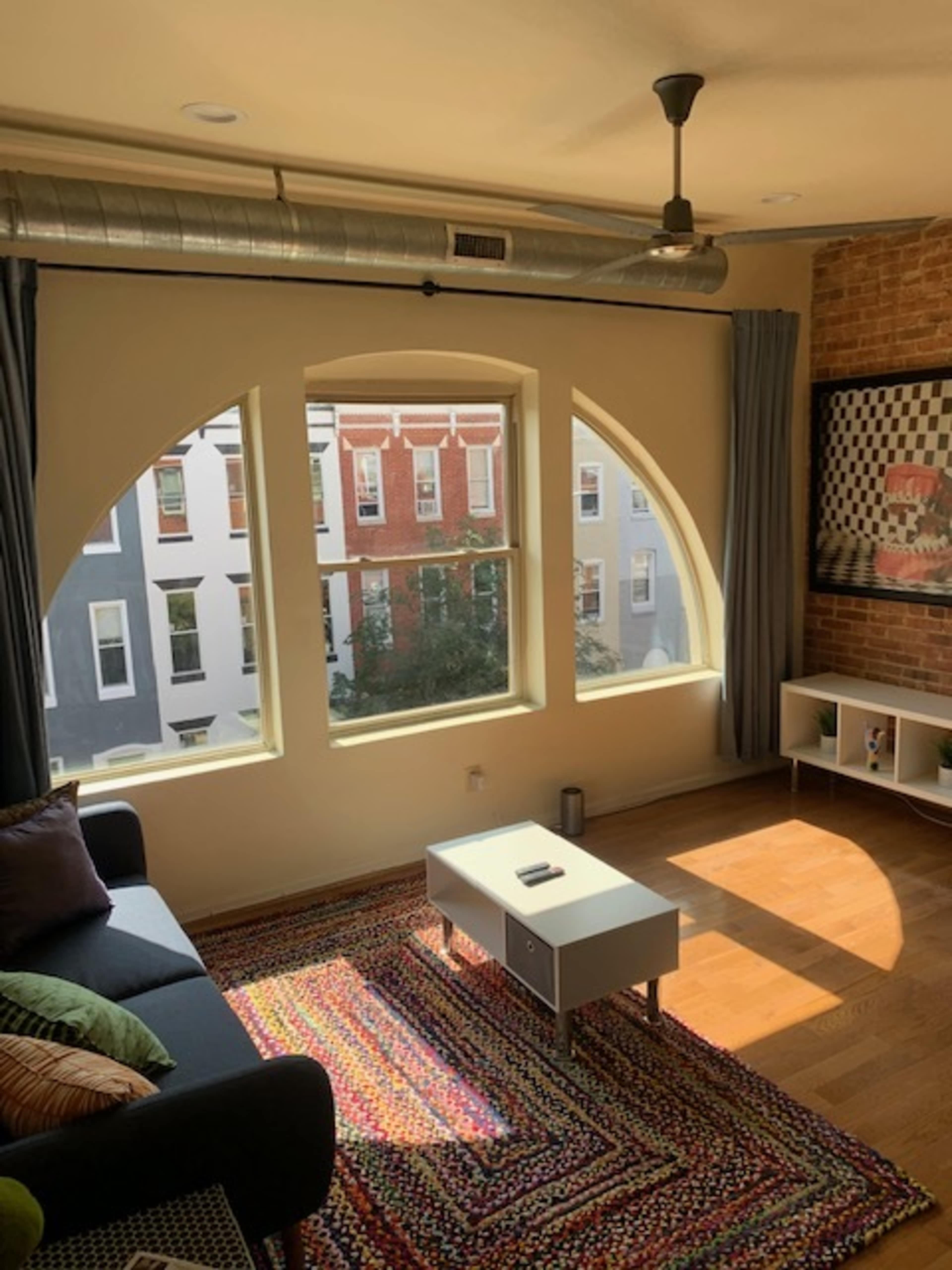 The image shows a living room with large arched windows, a dark sofa, a coffee table, and a colorful rug, set against a brick wall.