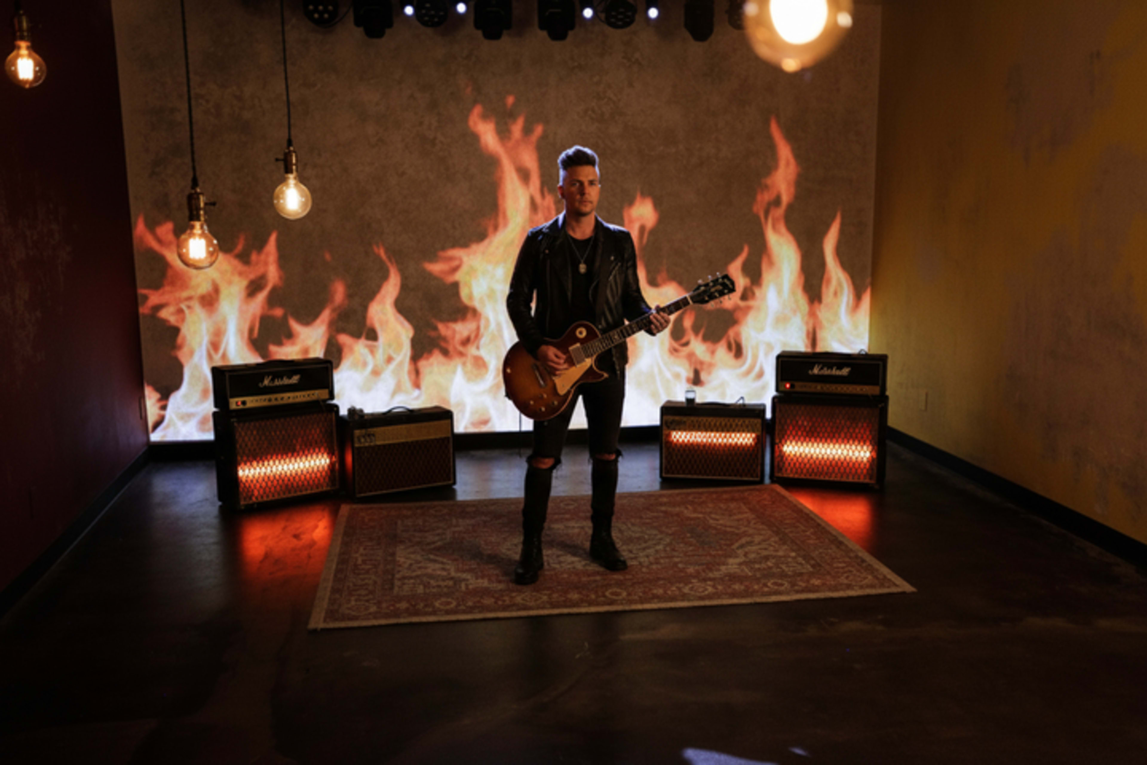 A musician stands on a rug in a dimly lit room with amplifiers and a backdrop of flames.