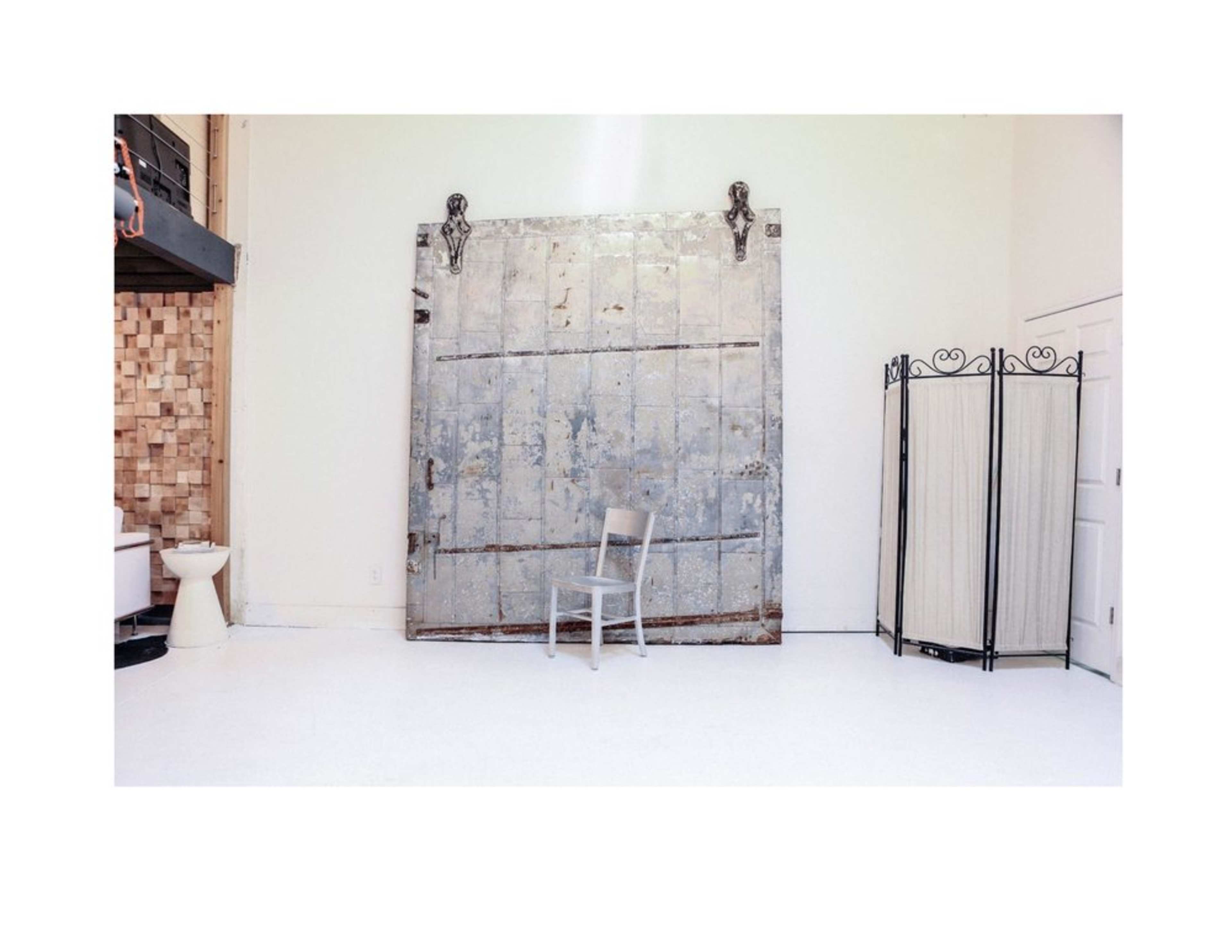 The image features a minimalist interior space with a large, weathered metal door, a single chair in front of it, and a folding screen nearby.
