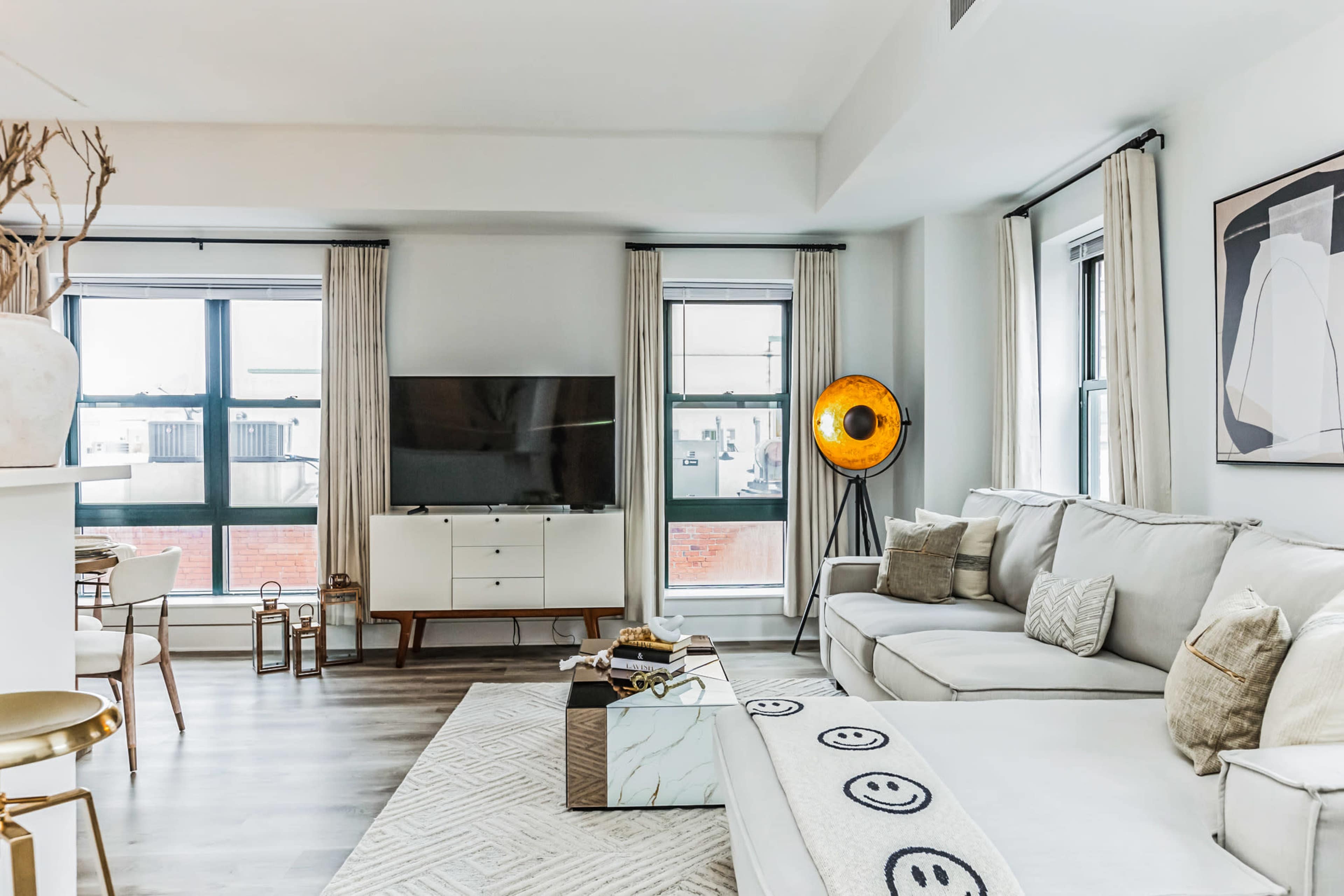 A modern living room featuring a sectional sofa, a television mounted on a white cabinet, and large windows with light curtains.