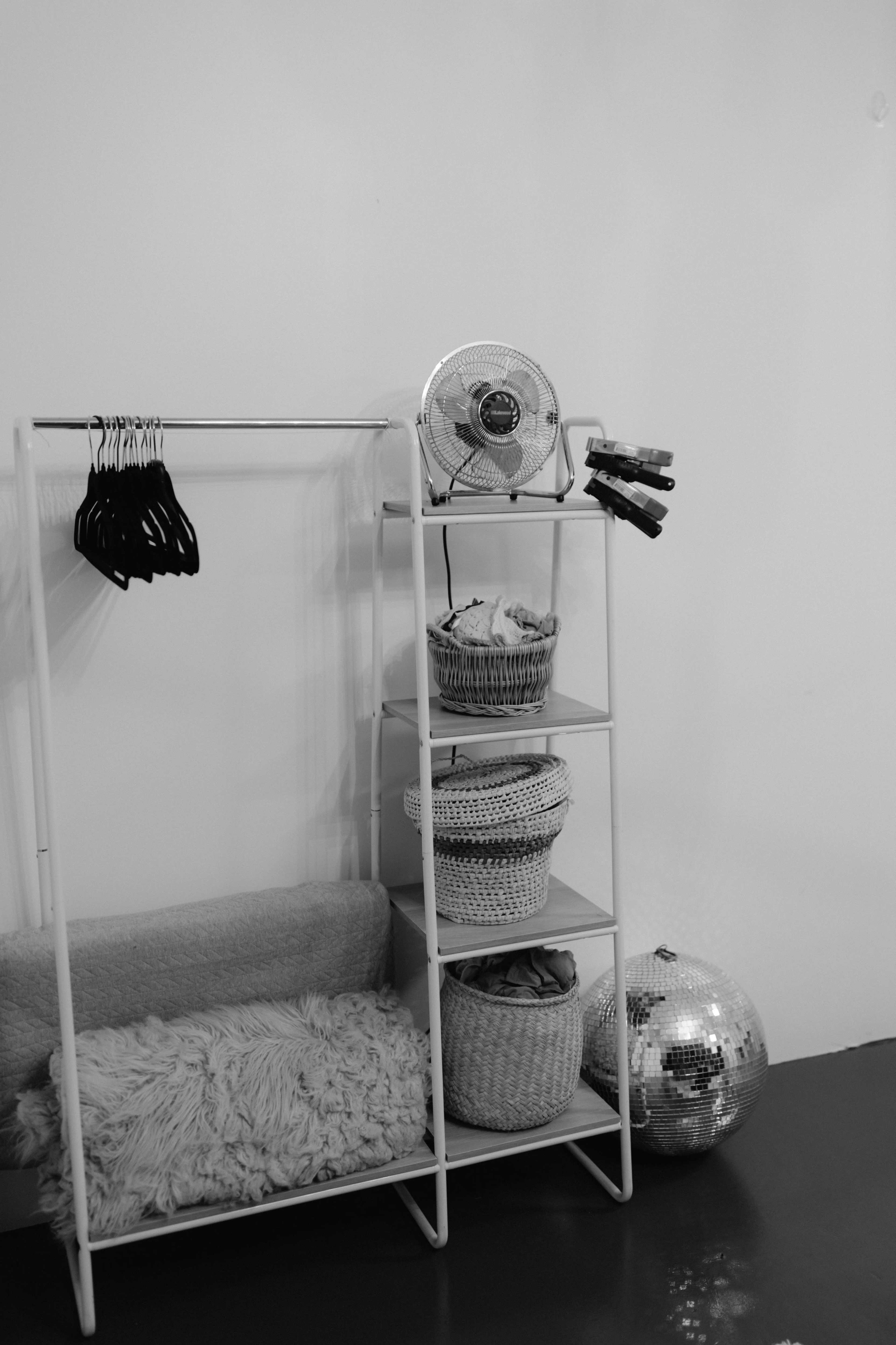 A metal clothing rack with hangers stands next to a shelf filled with woven baskets, a fan, and a disco ball on the floor.
