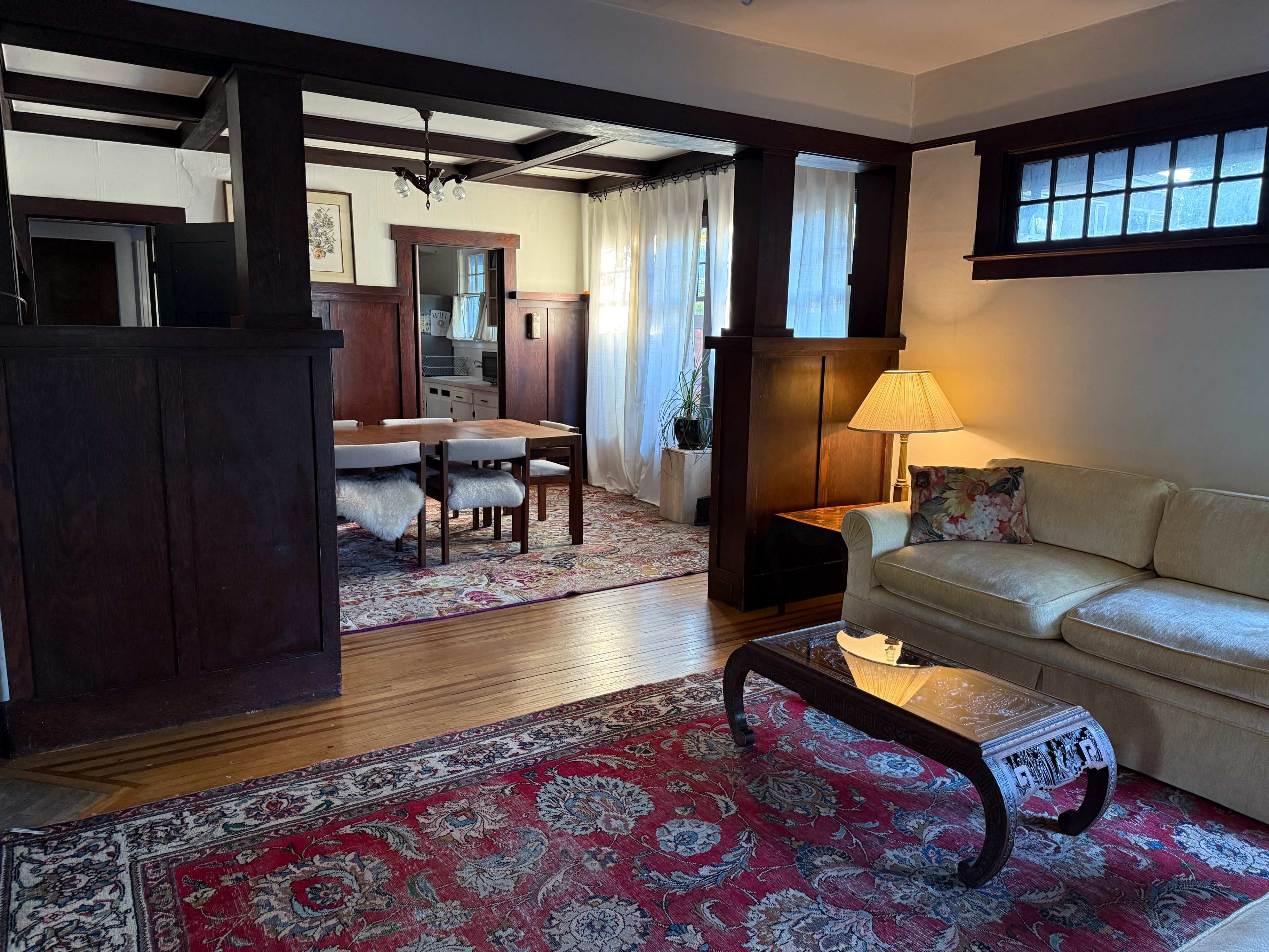 The image shows a living room with a tan sofa, a coffee table, and an adjacent dining area featuring a large table and chairs, all set under wooden beams and surrounded by large windows.