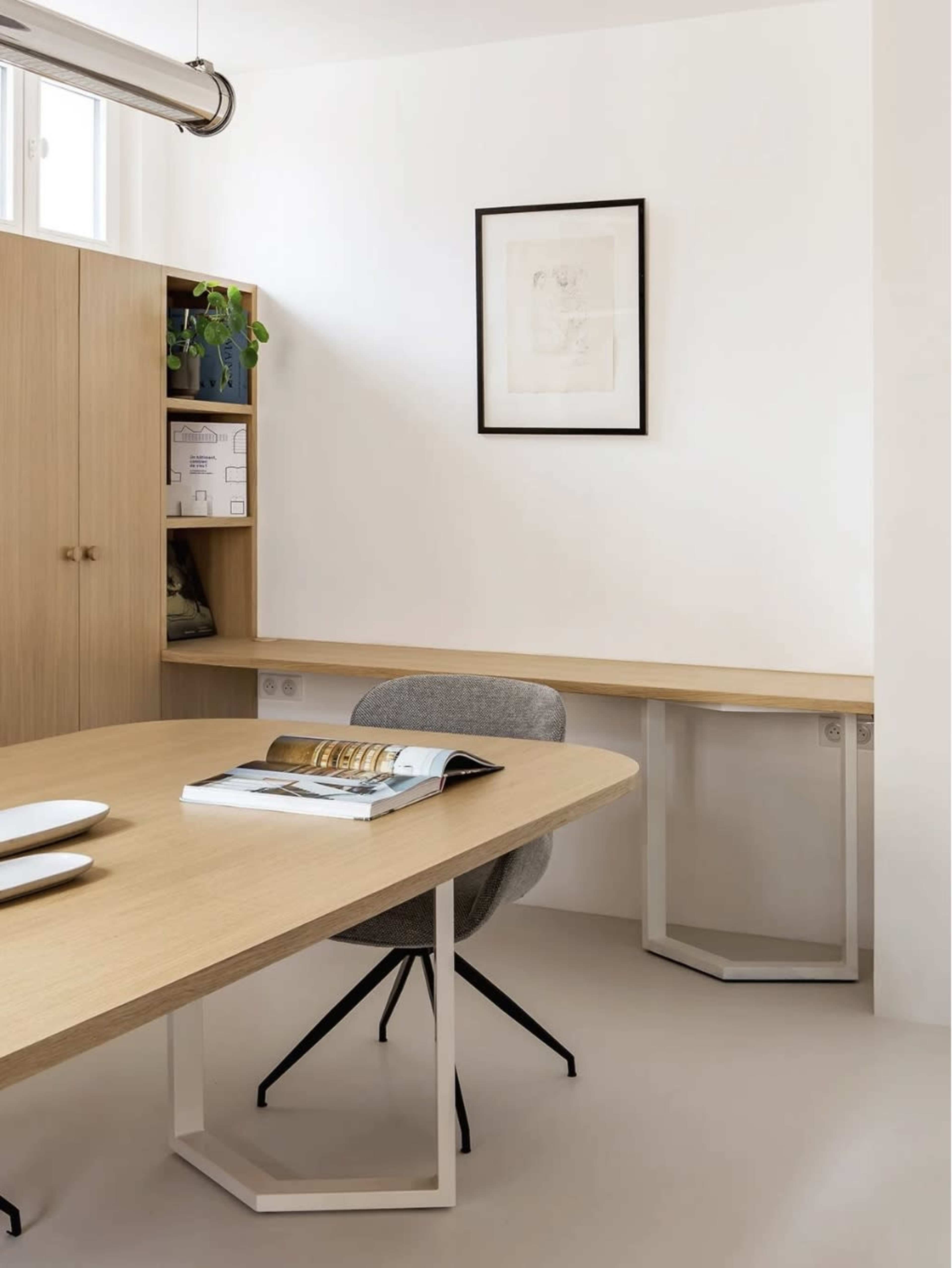 The image shows a minimalist office space featuring a wooden table, a gray chair, and a wall-mounted shelf containing books and a plant.