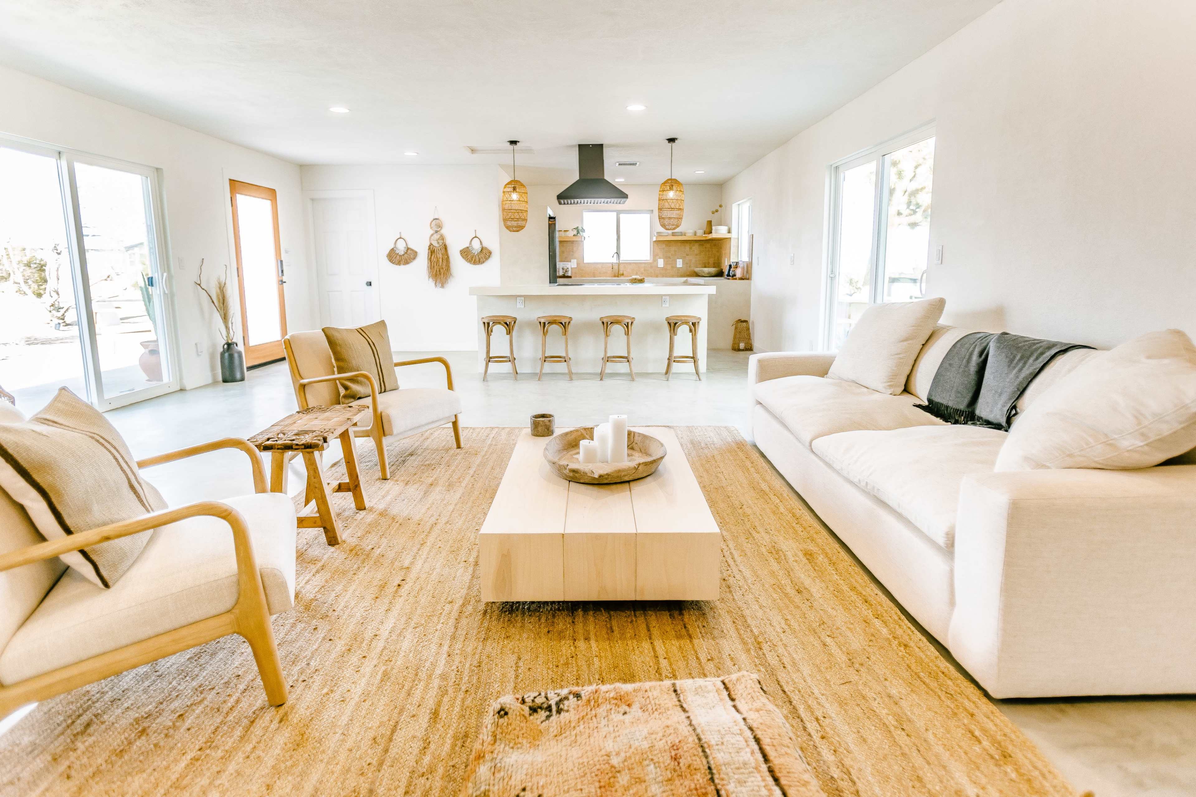 A modern living room with a white sofa, two wooden armchairs, a rectangular wooden coffee table, and a kitchen area visible in the background.