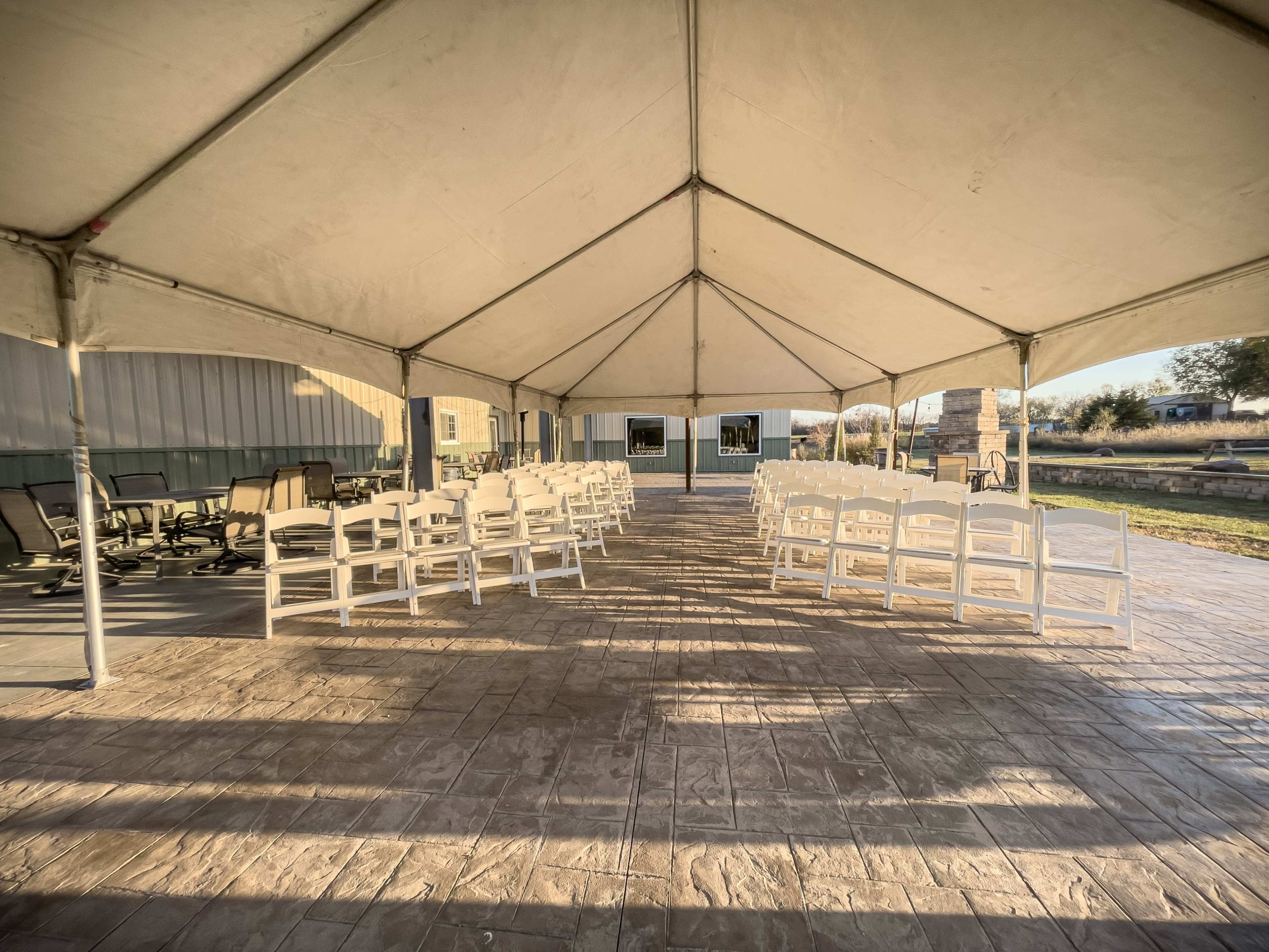 A large tent with white chairs arranged in rows on a patio, illuminated by sunlight.