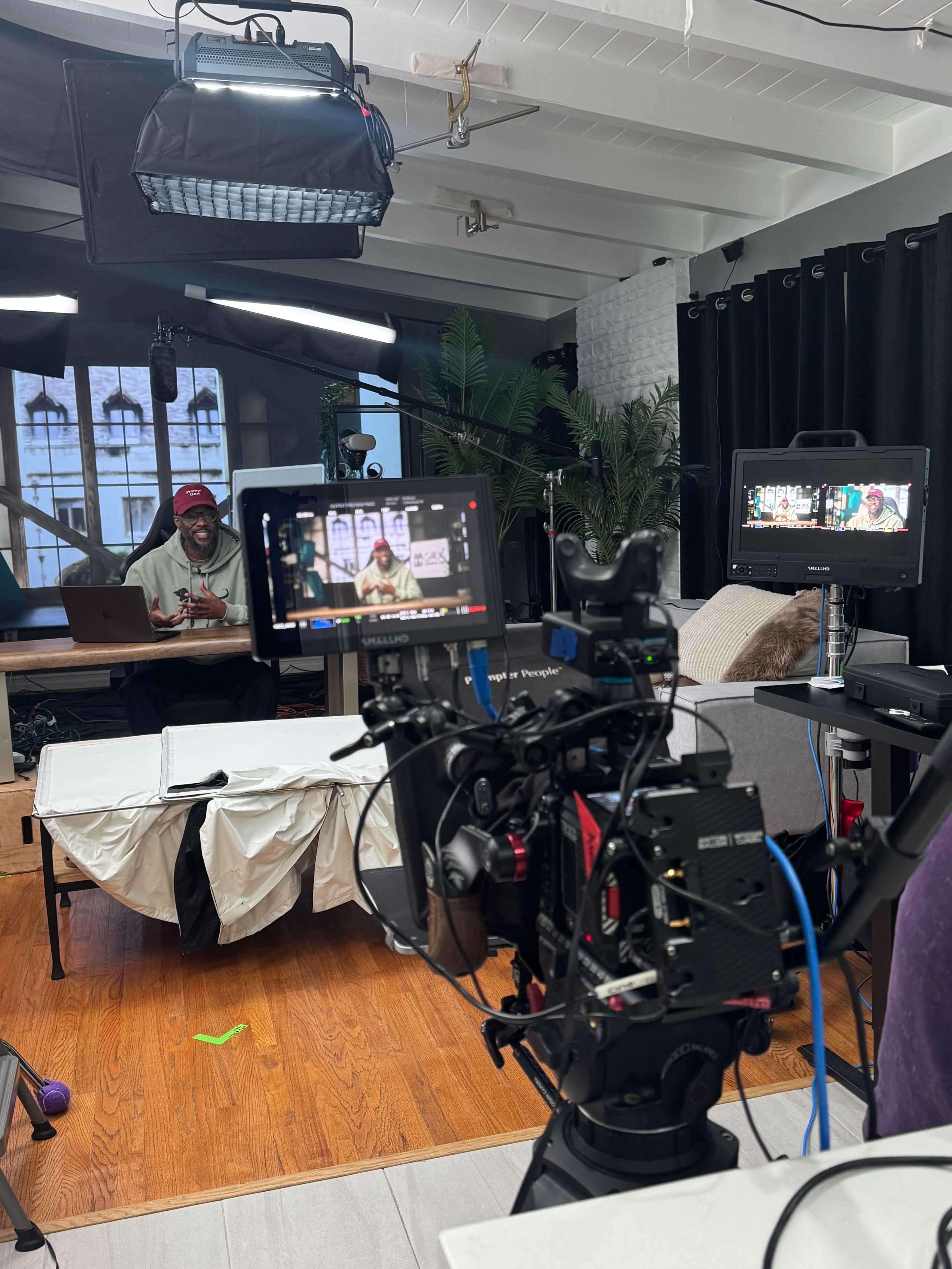 A camera setup is positioned in a studio space, capturing a person seated at a table, with additional monitors and lighting equipment visible.
