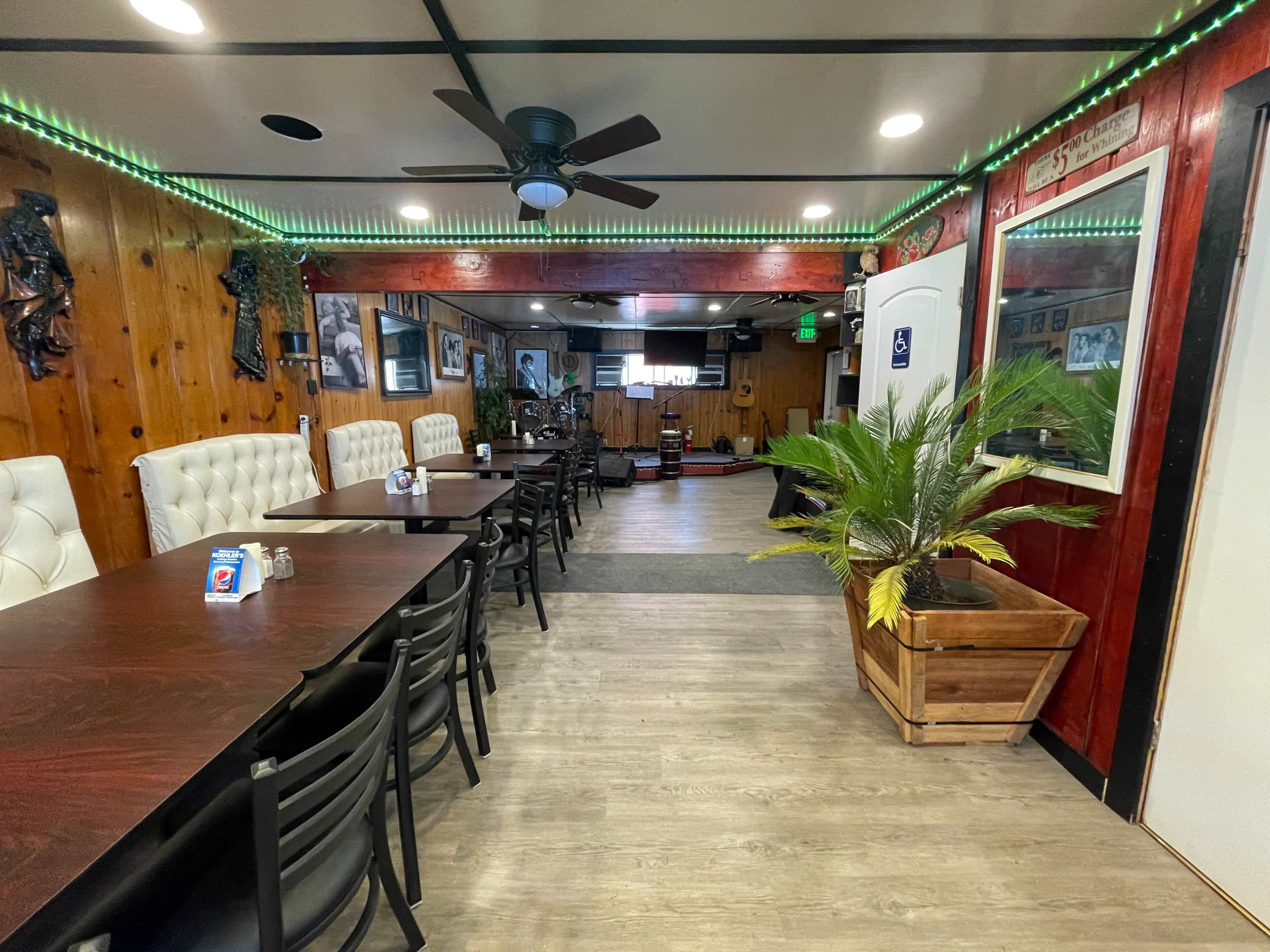 The interior of a restaurant features wooden paneling, tables with black chairs, and a potted plant near the entrance.