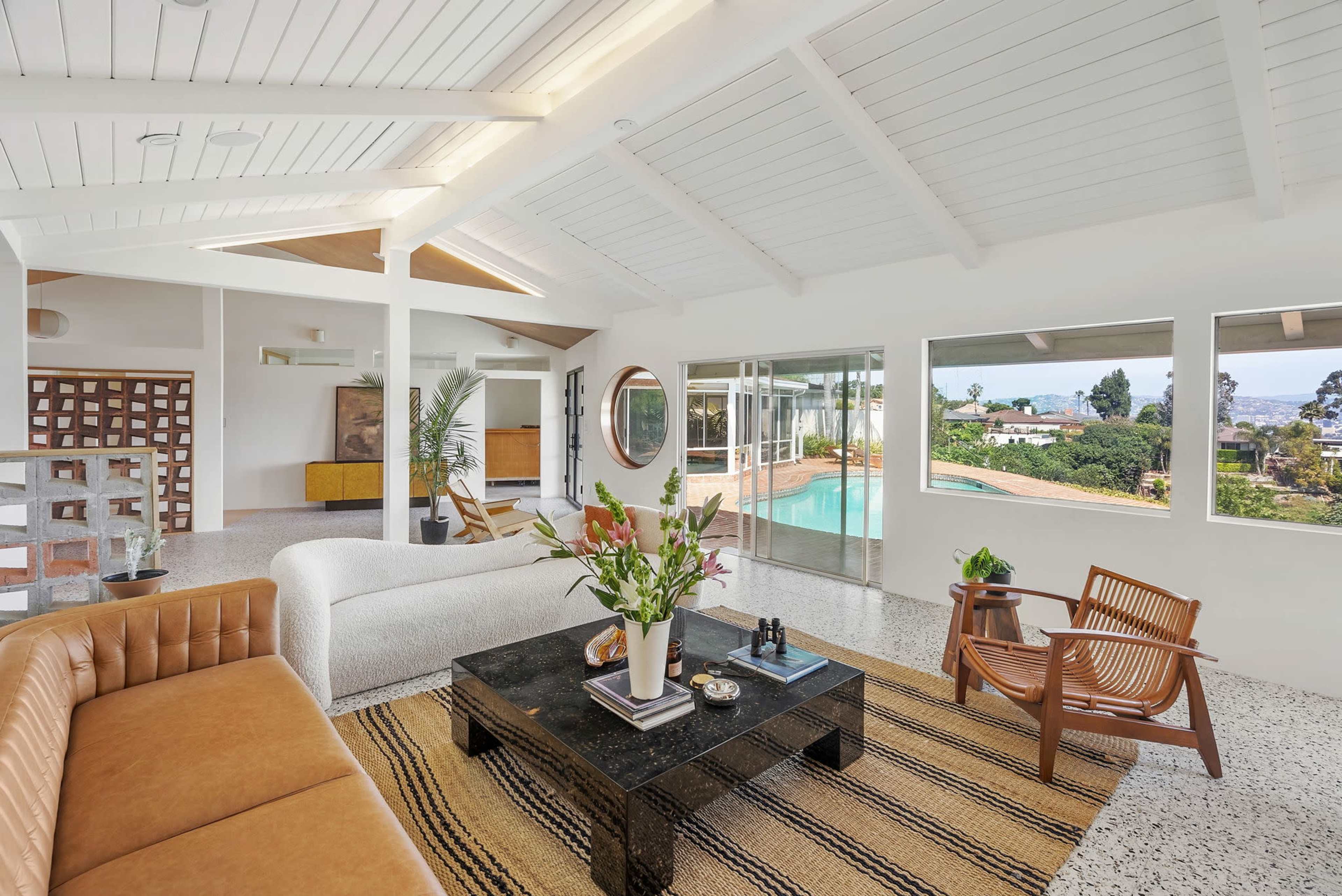 The image shows a modern living room with a beige sofa, a black coffee table, and large windows offering a view of an outdoor pool and garden.