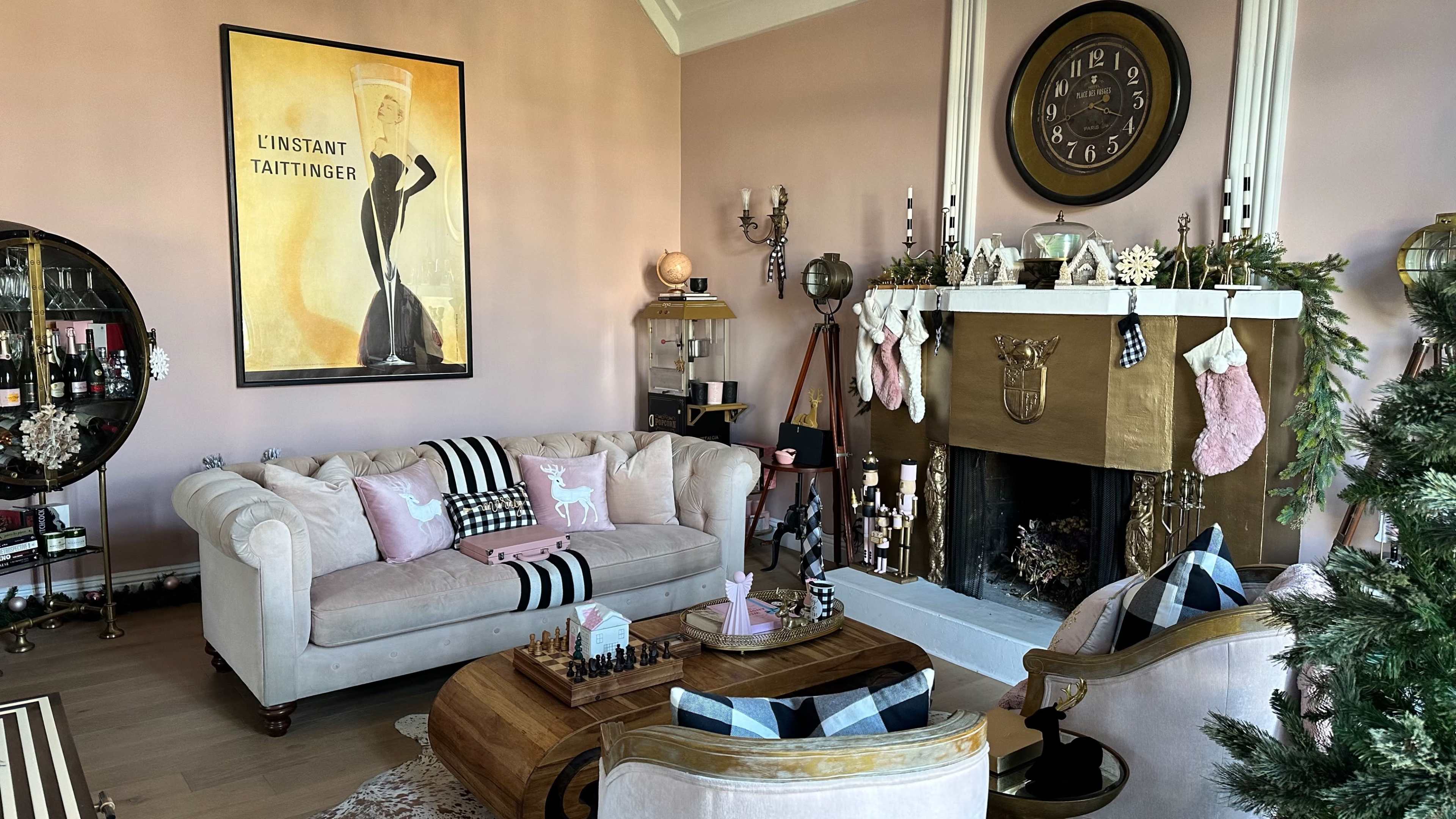 The image shows a pink-themed living room with a beige couch, a decorative fireplace adorned with stockings, and a large wall clock, all complemented by various pieces of furniture and decorations.