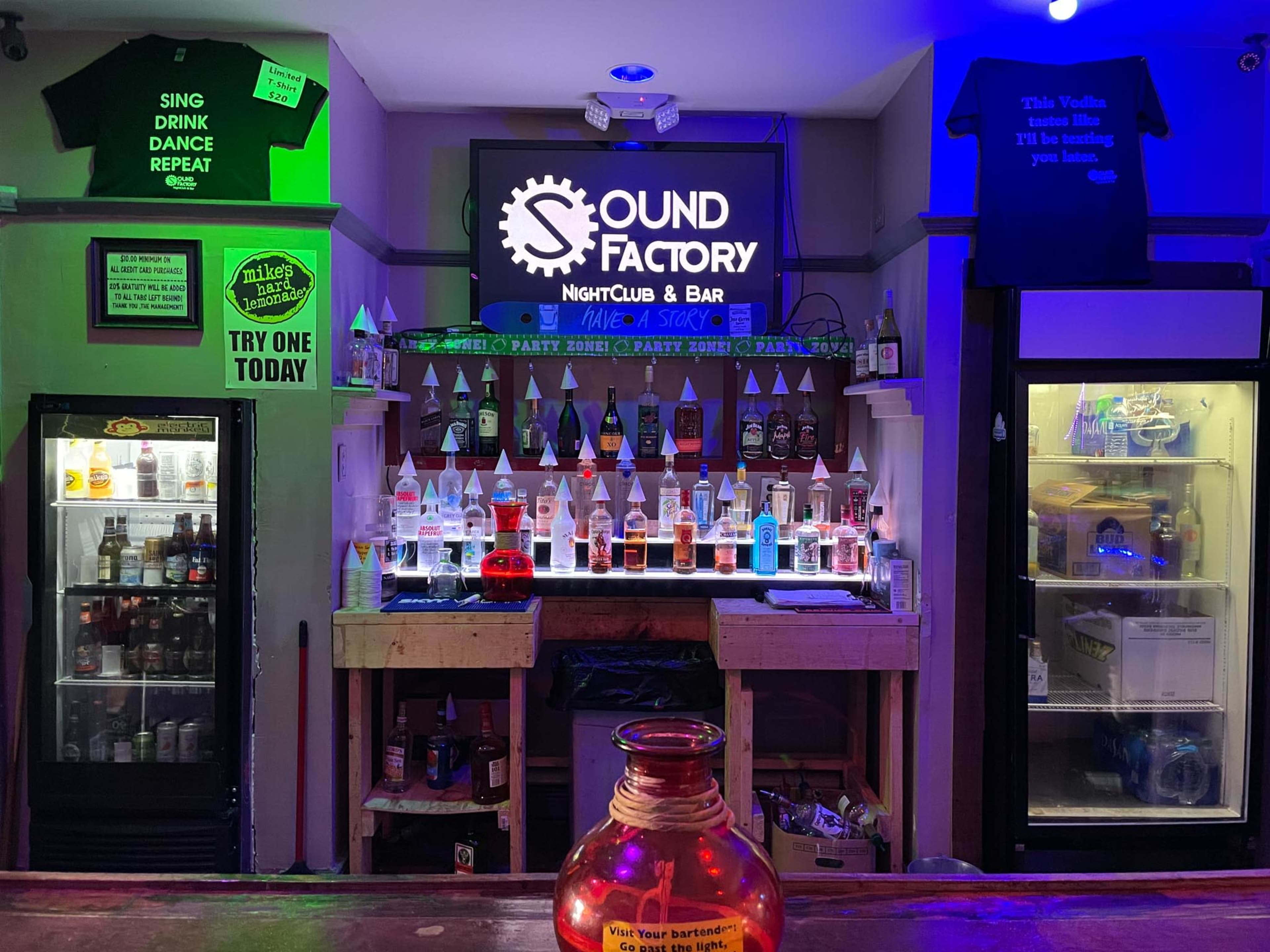 A bar area in a nightclub, featuring illuminated bottles of liquor on shelves, a neon sign reading "SOUND Factory NightClub & Bar," and various promotional items on display.