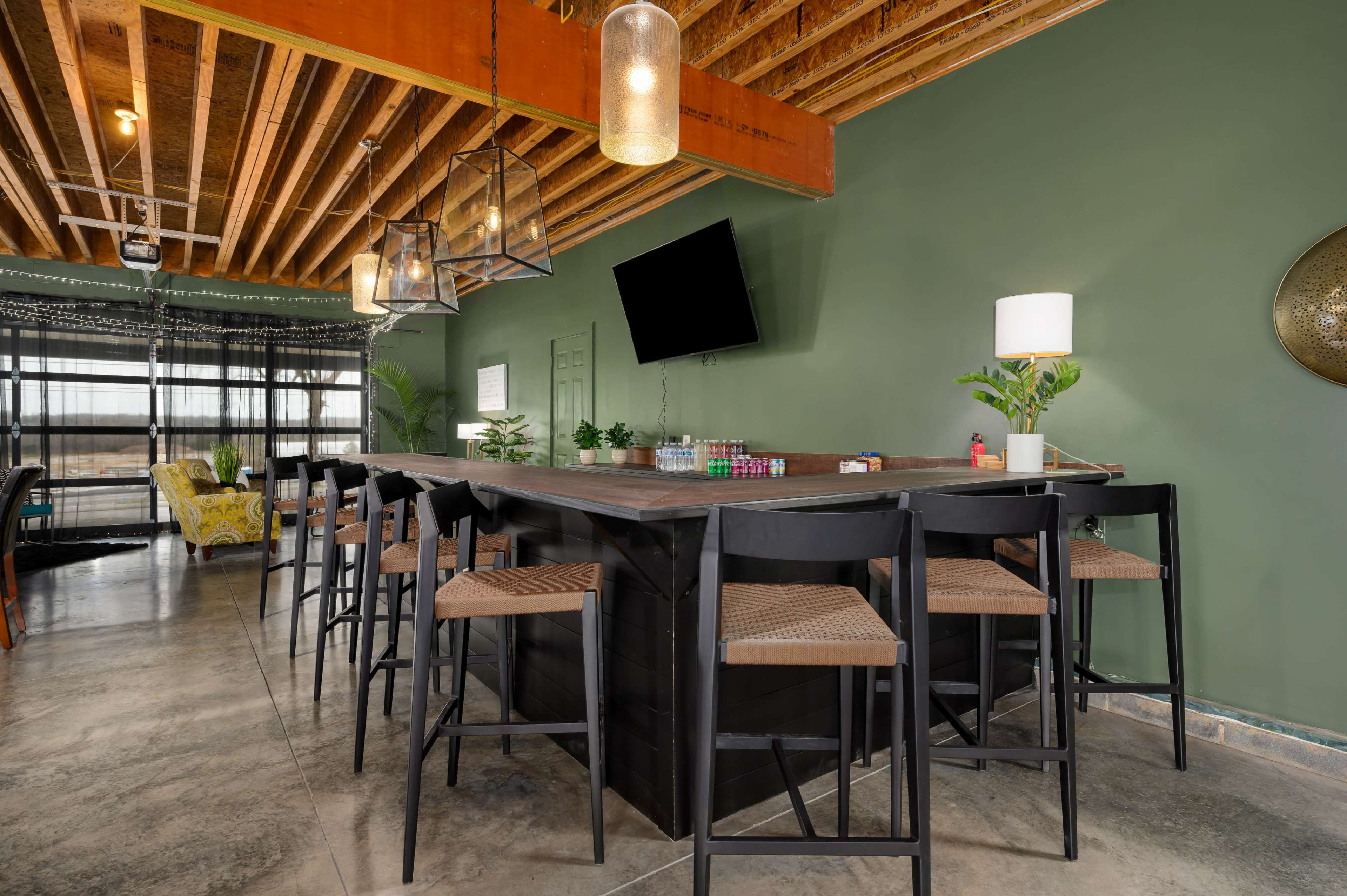 The image shows a modern bar area with high stools, a large table, and green walls, featuring hanging lights and a television on the wall.