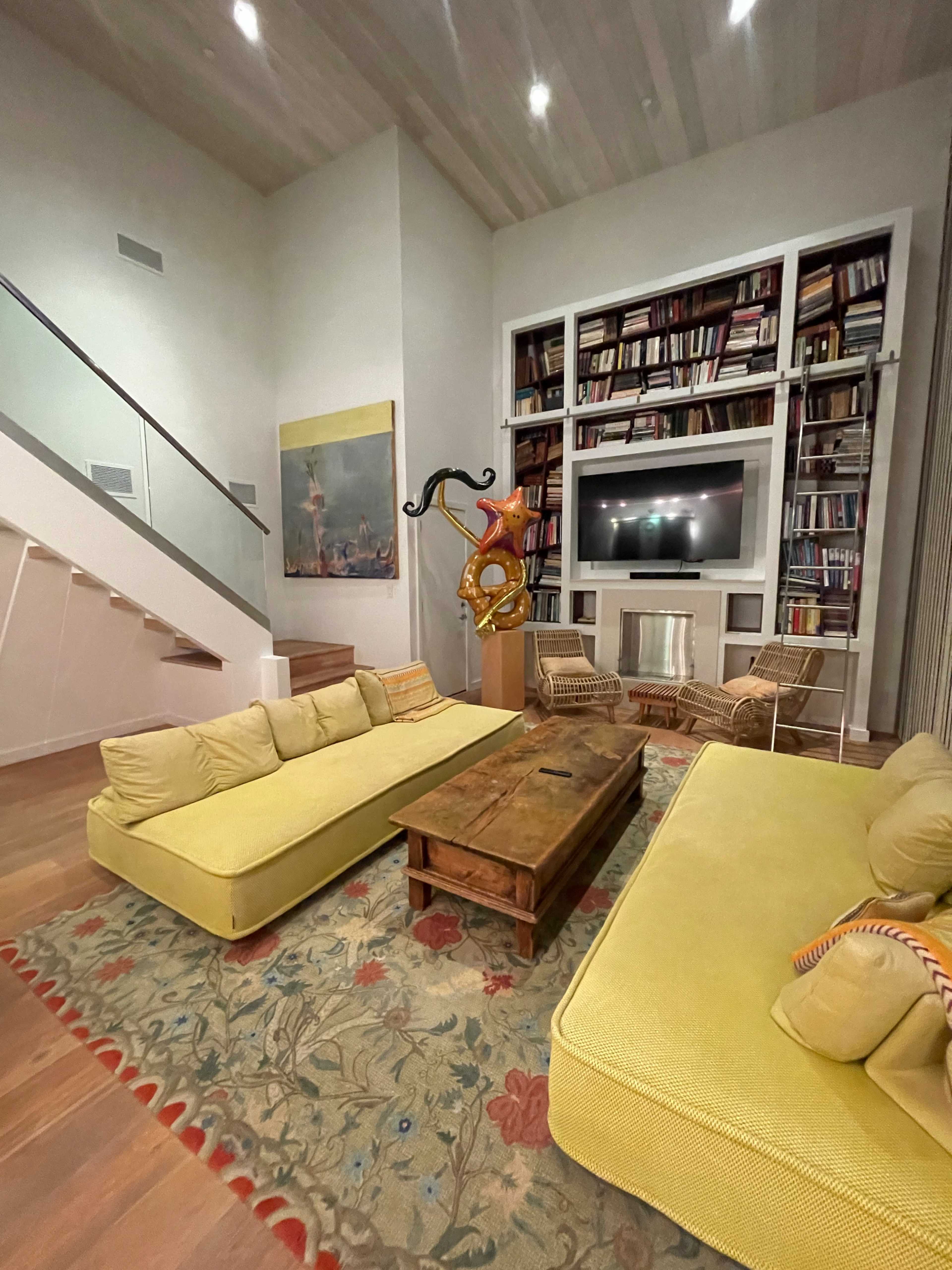 A modern living room features yellow sofas, a wooden coffee table, a colorful rug, and a large bookshelf filled with books.